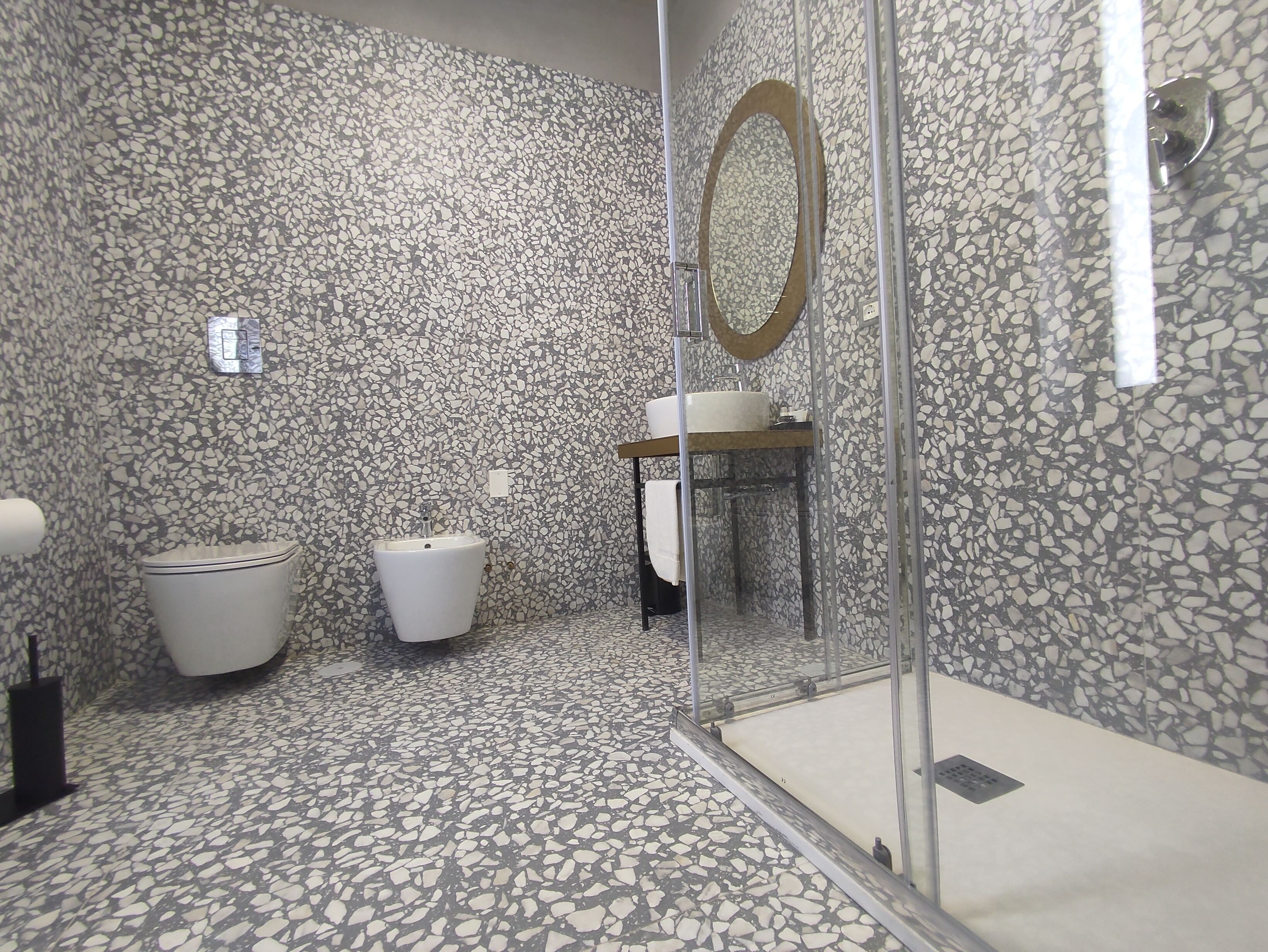 Une salle de bain moderne avec un sol en terrazzo, des toilettes, un lavabo et une douche séparée par une paroi de verre.