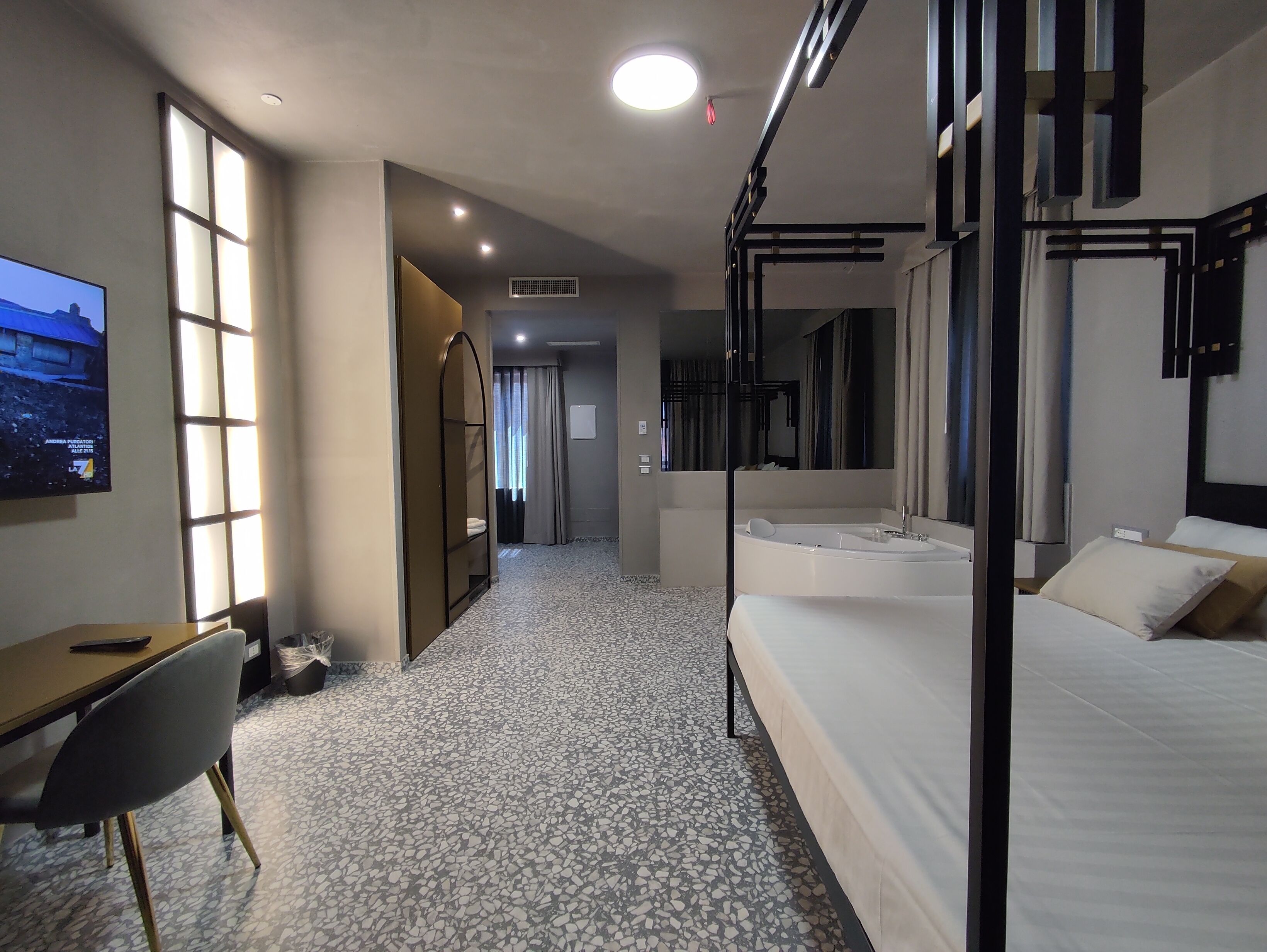 Habitación de hotel con cama, escritorio, silla, un espejo grande y televisor en la pared.