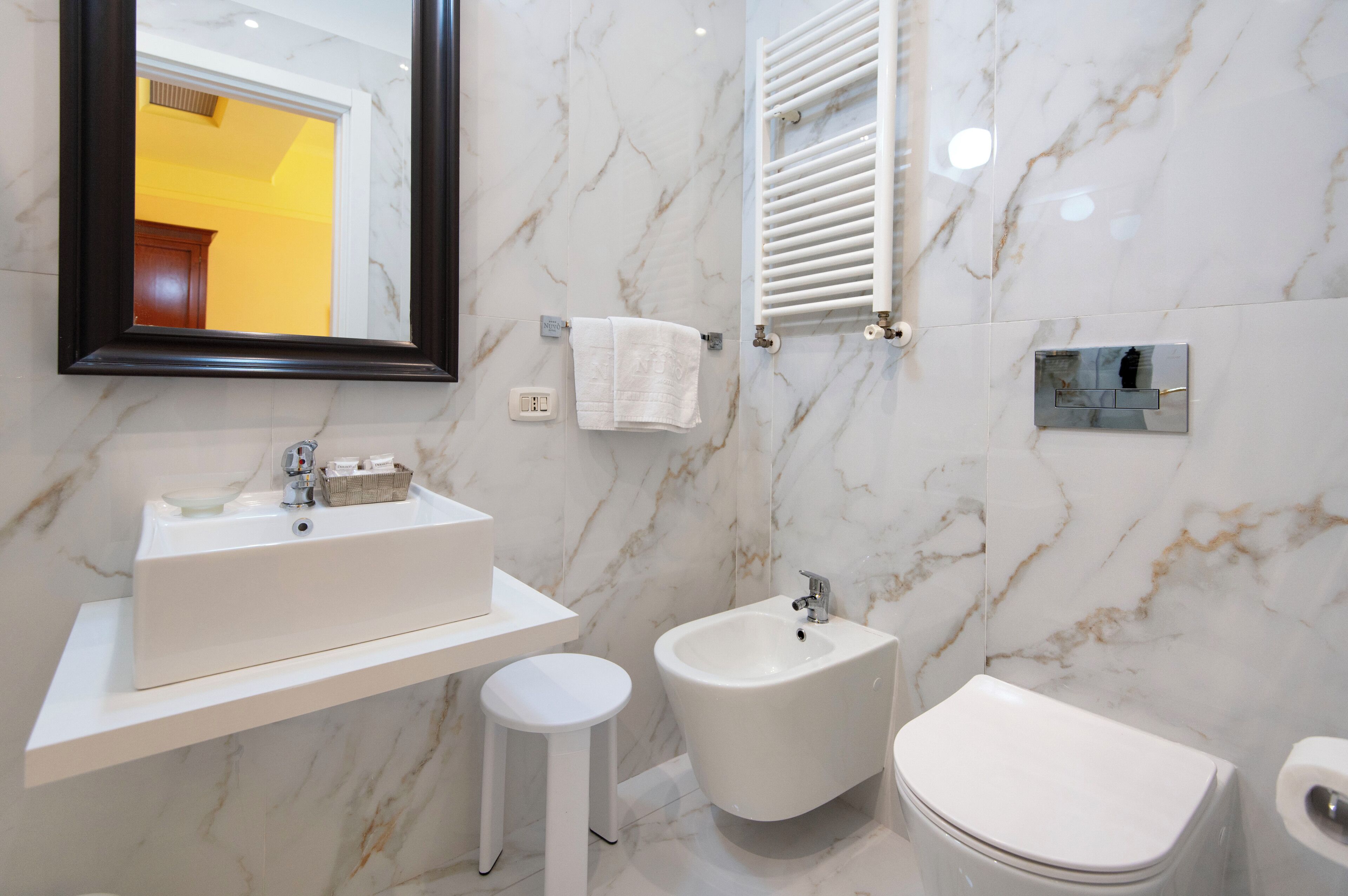 Ein modernes Badezimmer mit einem weißen Waschbecken, einem Bidet und einer Toilette, Marmorwänden und einem Spiegel mit schwarzem Rahmen.