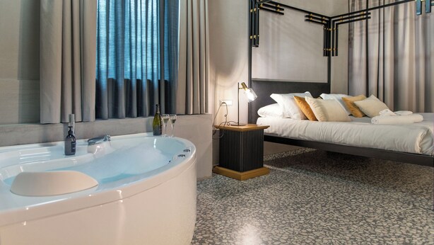 Quarto de hotel moderno com uma banheira grande, cama com roupa de cama branca e bege, e um espelho com moldura preta.