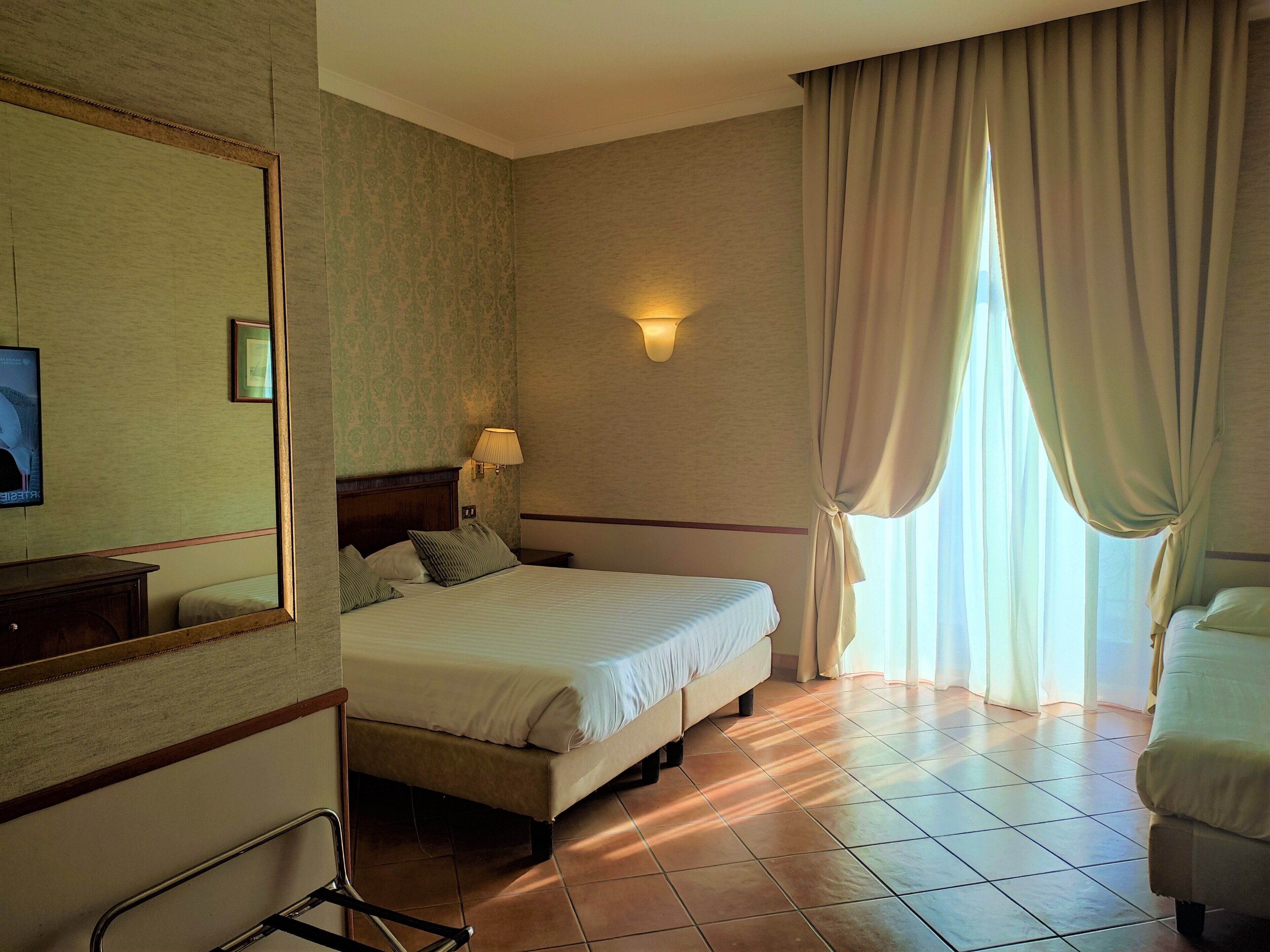A hotel room with a bed, a bedside table, a lamp, a chair, a large mirror, and a window with curtains.