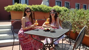 Duas mulheres sentadas numa mesa ao ar livre com bebidas, rodeadas por plantas em vasos e um edifício ao fundo.