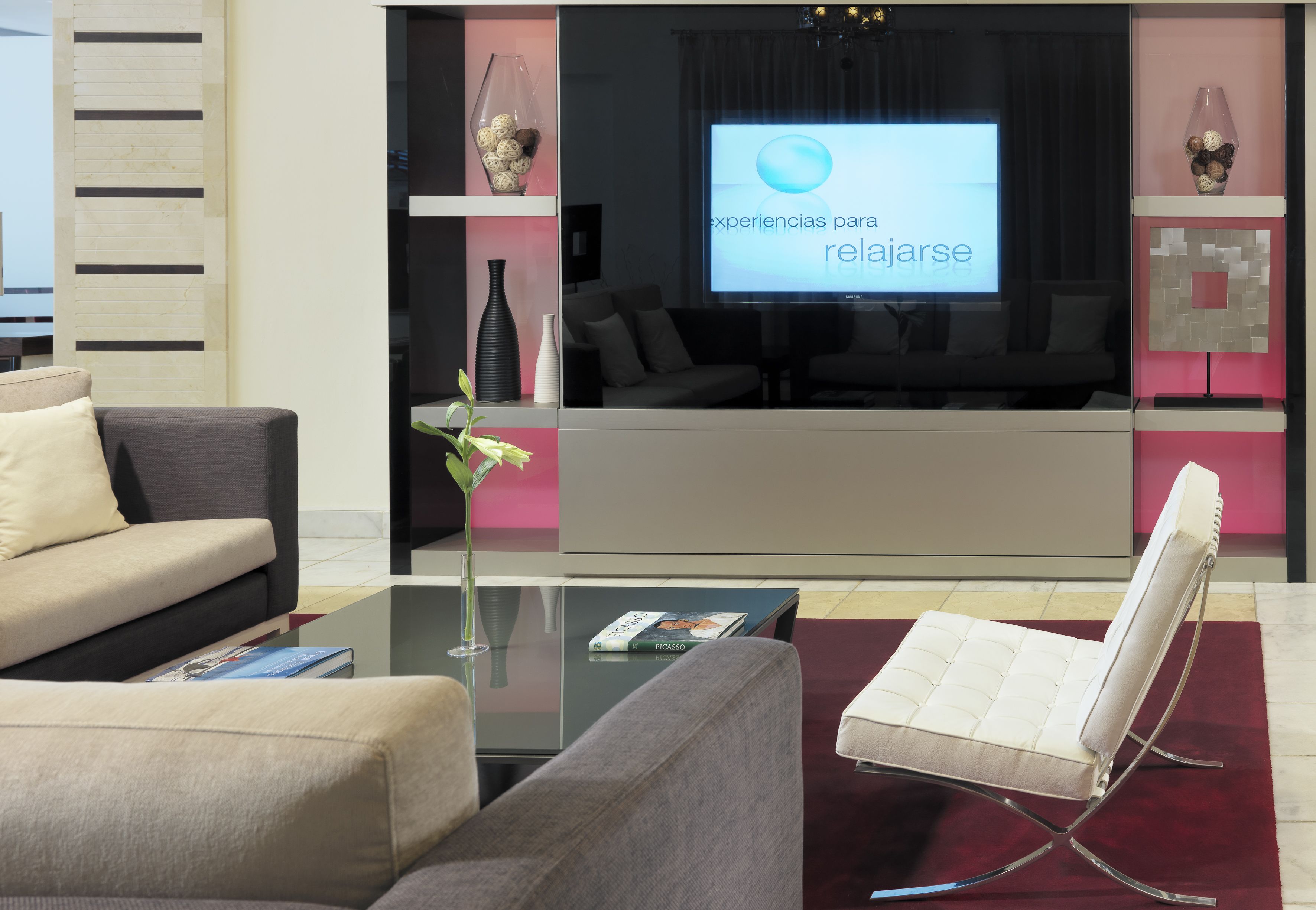 Un salón moderno con un sofá, una silla blanca, una mesa de centro de vidrio y una pantalla de televisión mostrando 'experiencias para relajarse'.