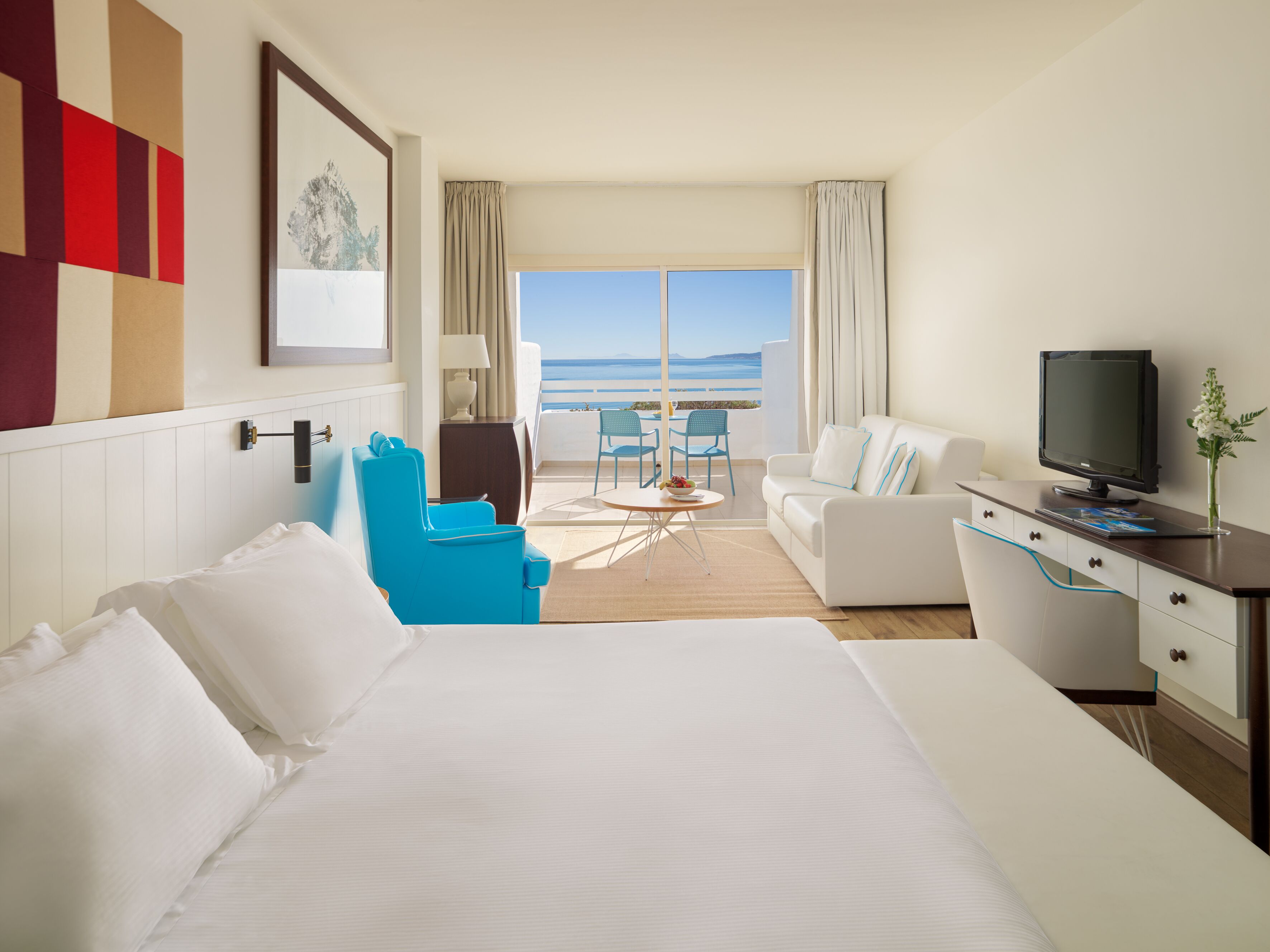 Quarto de hotel com cama, uma escrivaninha com televisão, área de estar com cadeiras e vista para o mar.