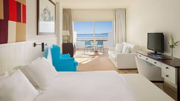 Quarto de hotel com cama, uma escrivaninha com televisão, área de estar com cadeiras e vista para o mar.