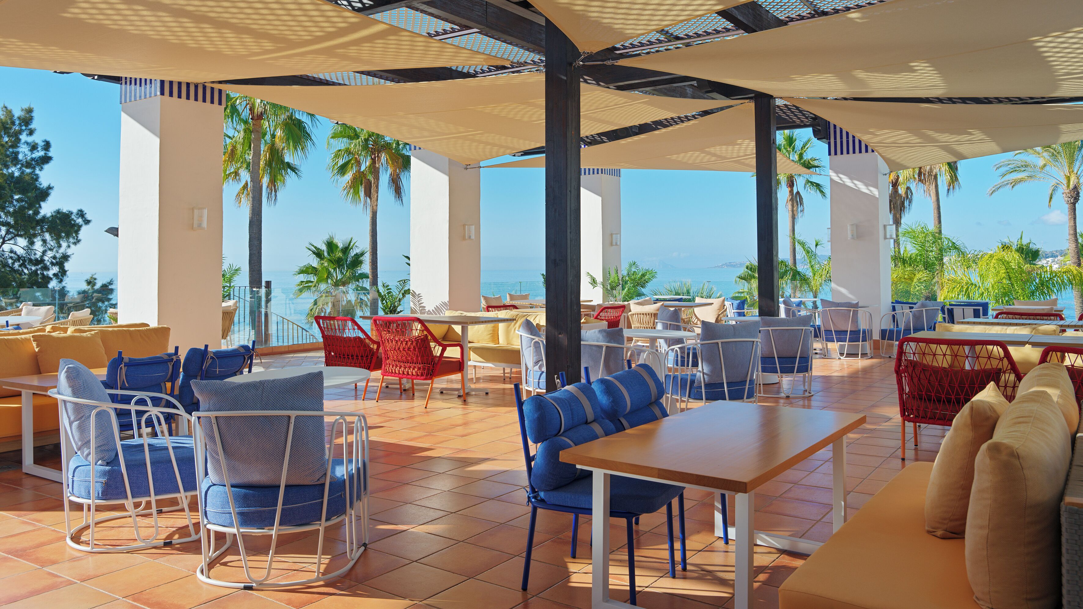 A terrace with outdoor seating, palm trees, and a view of the sea.
