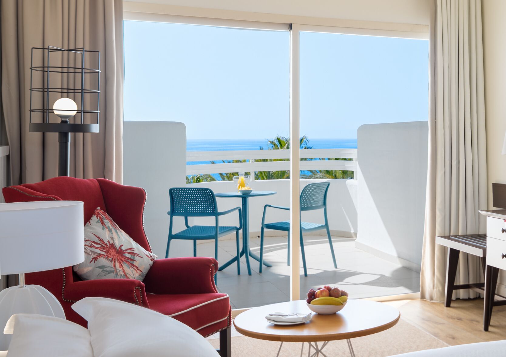 A room with a red sofa, white armchairs, a small table with a bowl of fruit, and a balcony with a view of the ocean.