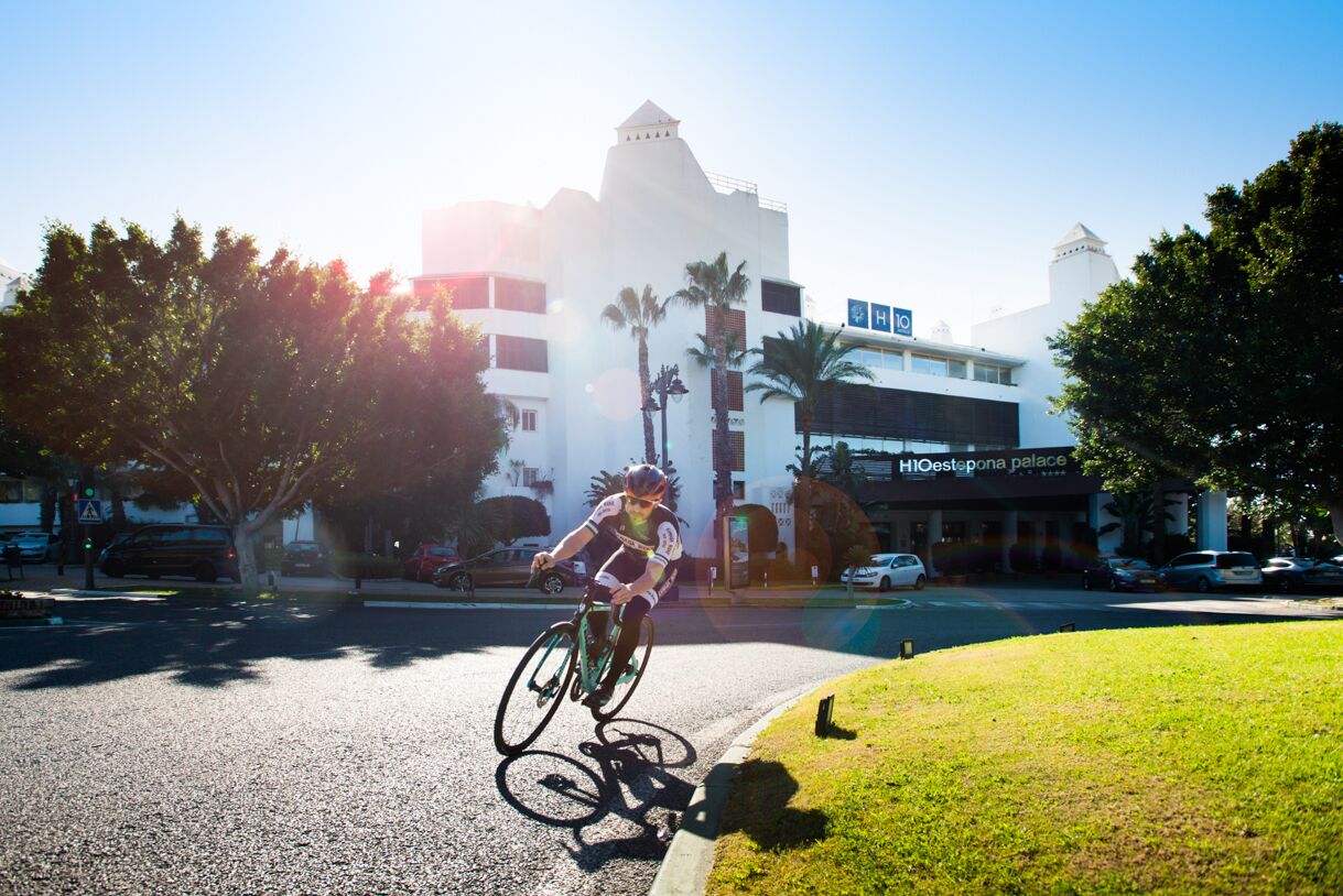 Un ciclista pedaleando por una carretera, con un edificio con la leyenda "HIOestepona palace" al fondo.