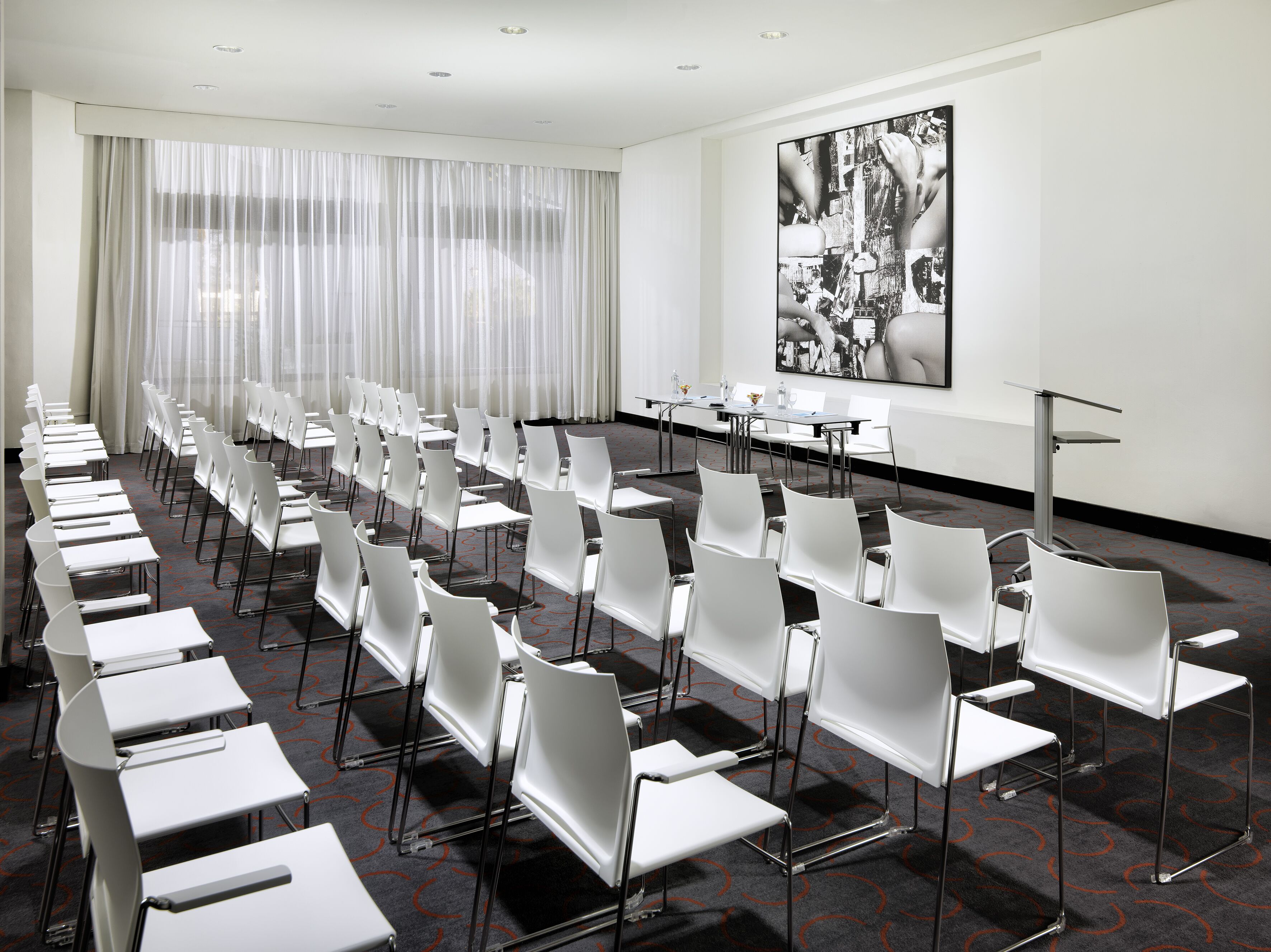 Un salón de conferencias con filas de sillas y mesas blancas, un amplio ventanal con cortinas translúcidas y una obra de arte blanco y negro en la pared.