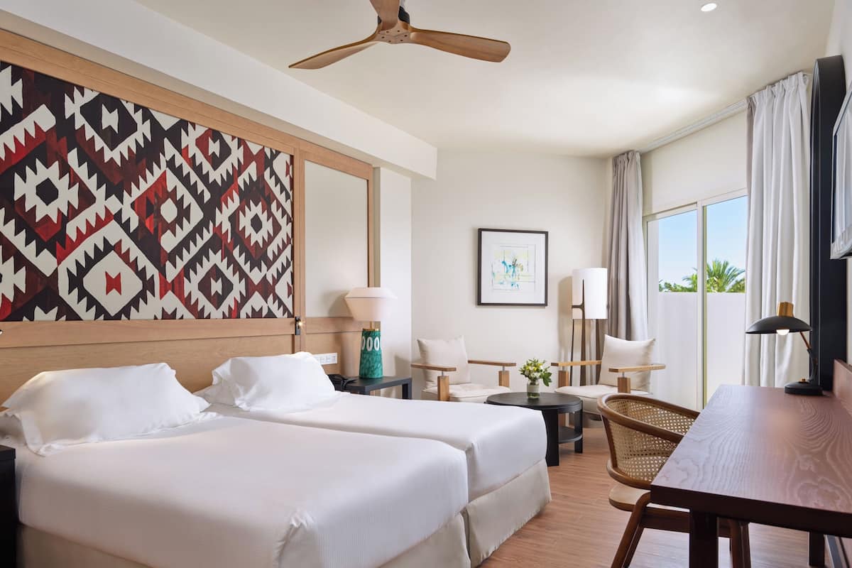 A hotel room with two beds, a ceiling fan, a large window with curtains, a desk, and a chair.