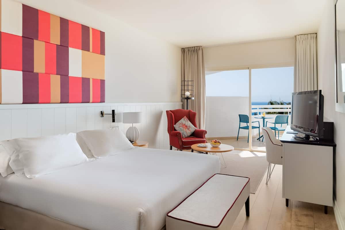 A hotel room with a large bed, a red armchair, a small round table with a bowl of fruit, a television on a cabinet, and a balcony with a view of the sea.