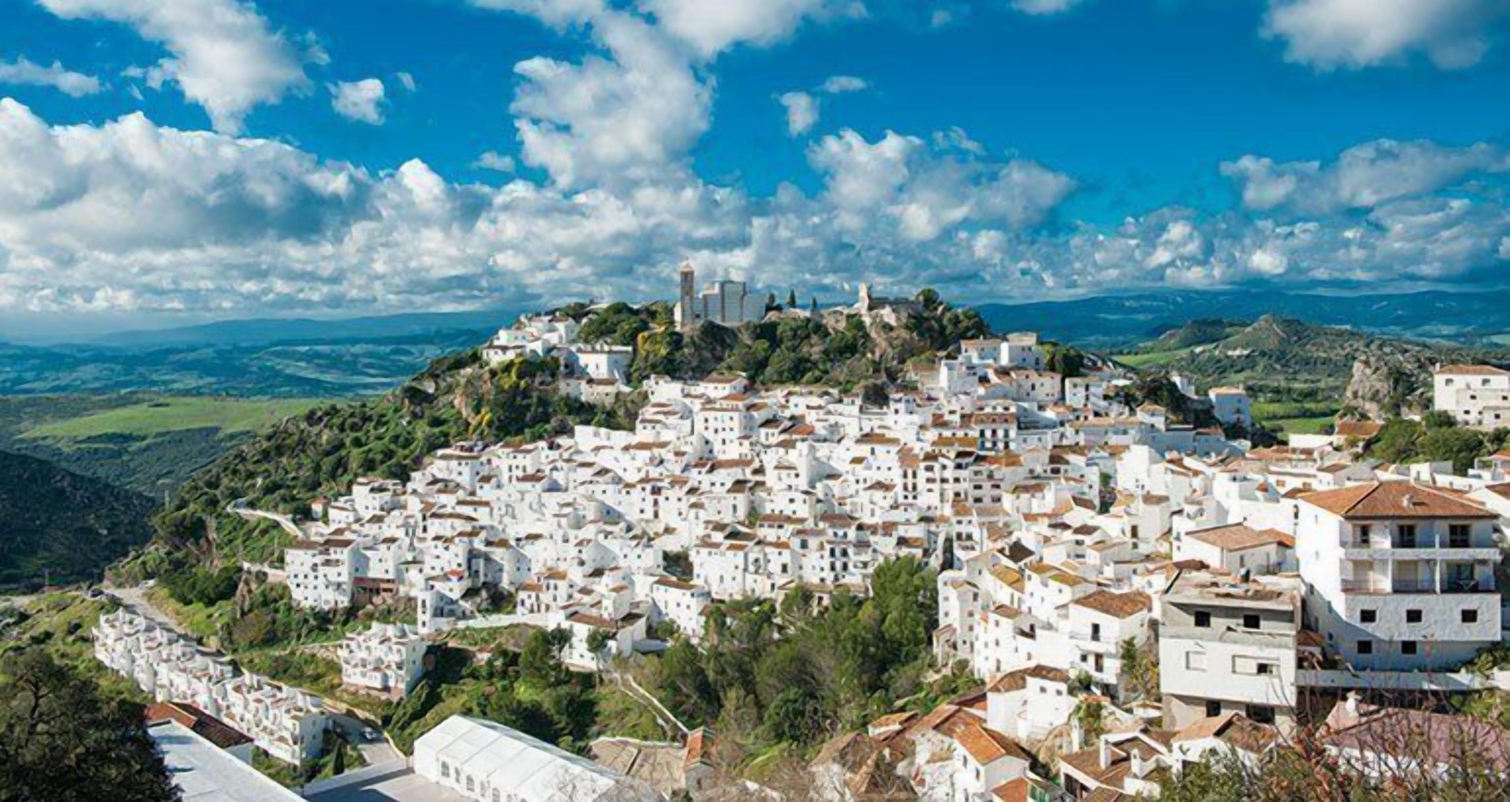 Un pueblo costero con edificios blancos, una torre destacada y vistas al mar y las colinas.