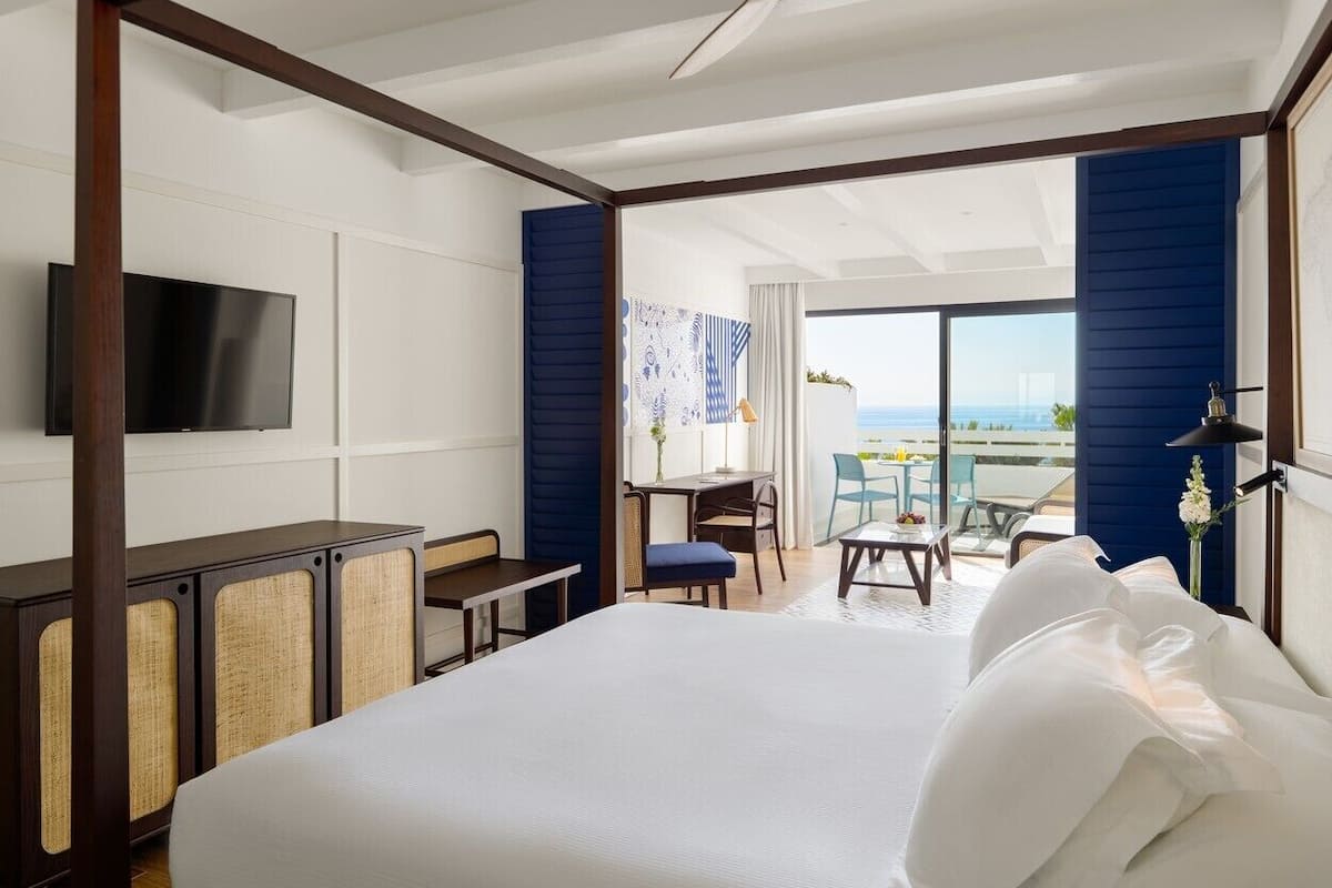 A modern hotel room with a large bed, a desk, and a balcony with a view of the sea.
