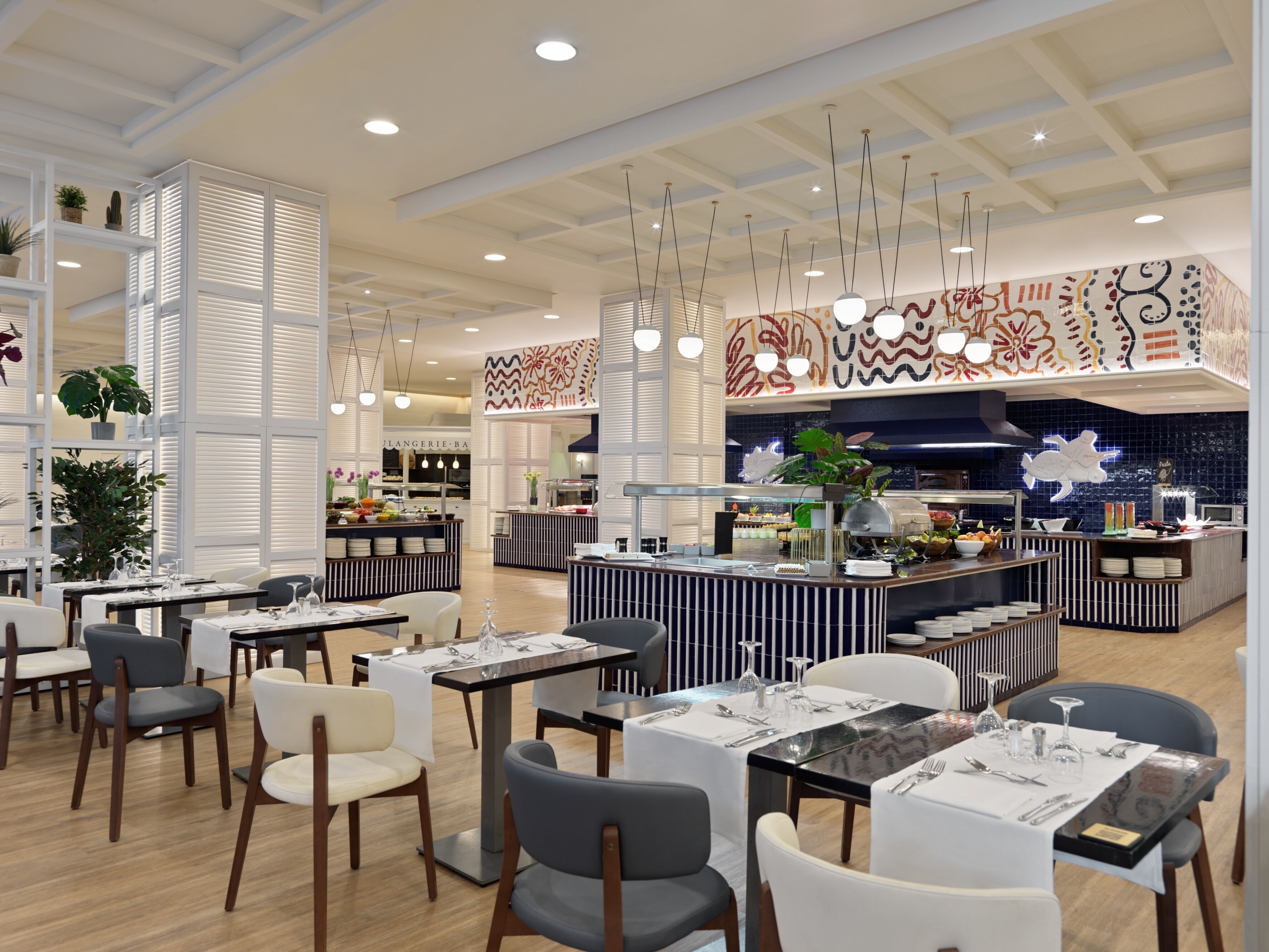 A modern restaurant with high ceilings, pendant lighting, and a central bar area. Tables are neatly arranged with chairs, and the decor includes patterned wall art and potted plants.