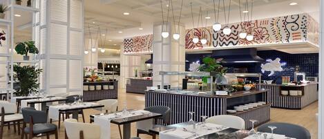 A modern restaurant with high ceilings, pendant lighting, and a central bar area. Tables are neatly arranged with chairs, and the decor includes patterned wall art and potted plants.