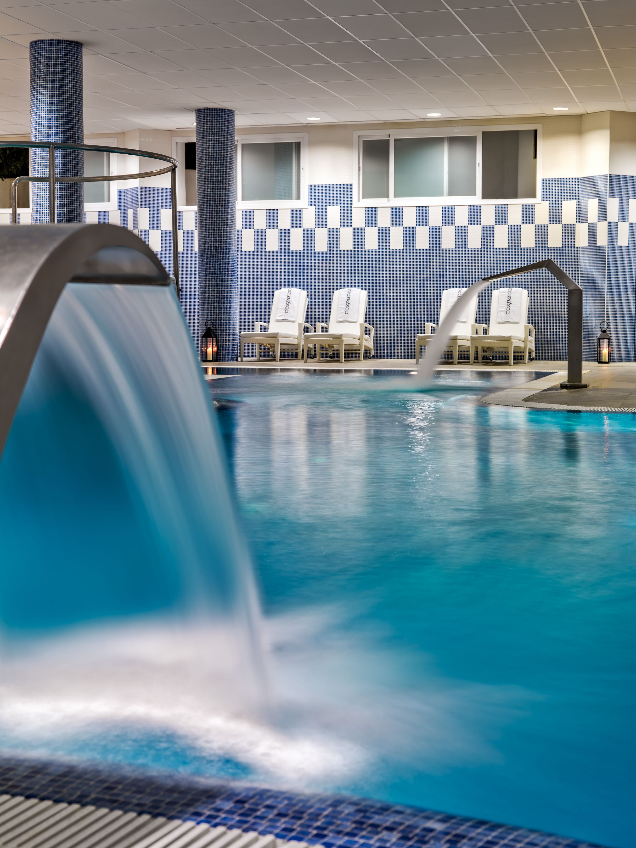 A swimming pool with a water feature, lounge chairs, and a tiled wall with a checkered pattern.