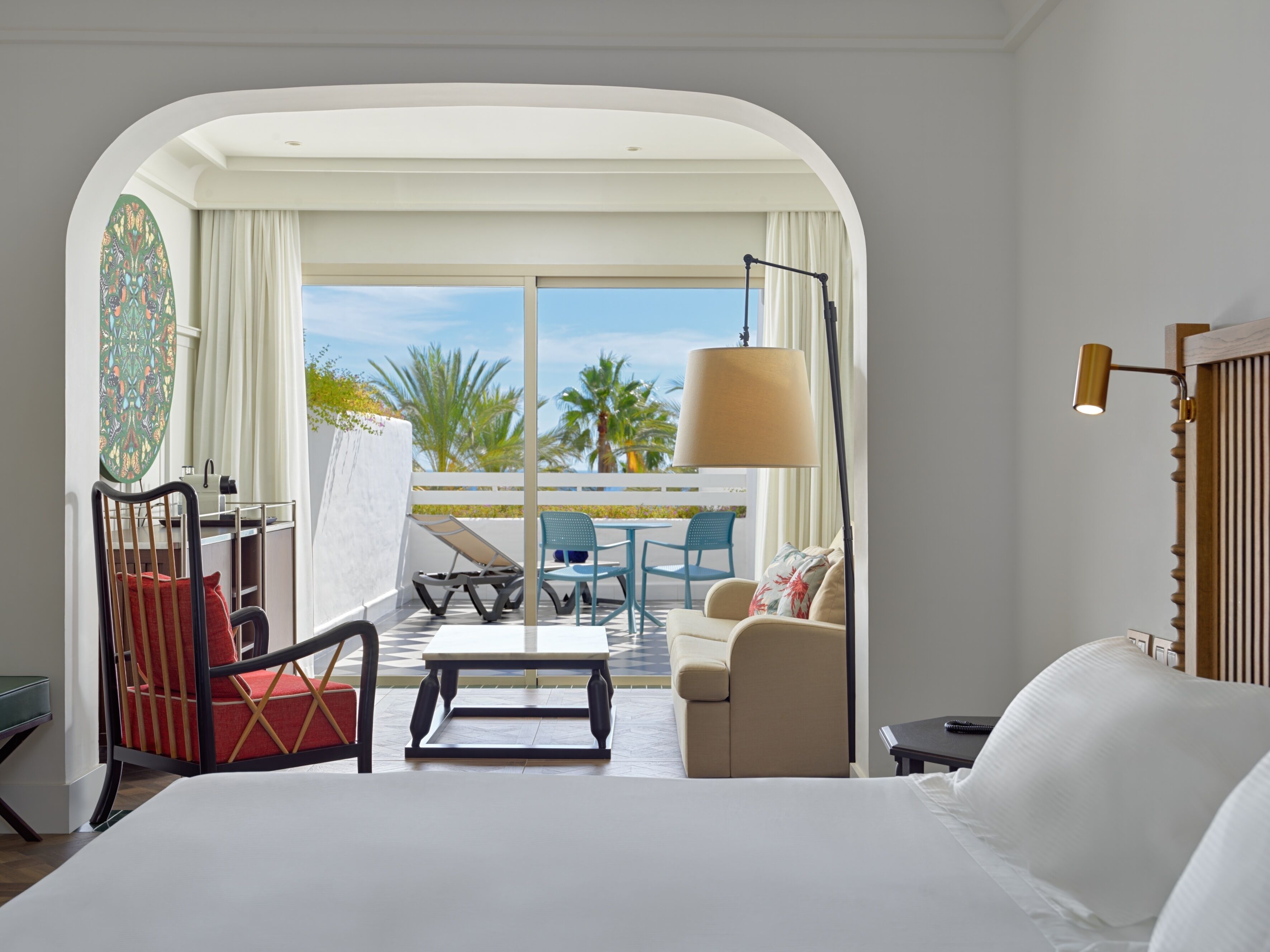 A hotel room with a large bed, a desk, a chair, a sofa, and a balcony with a view of palm trees.