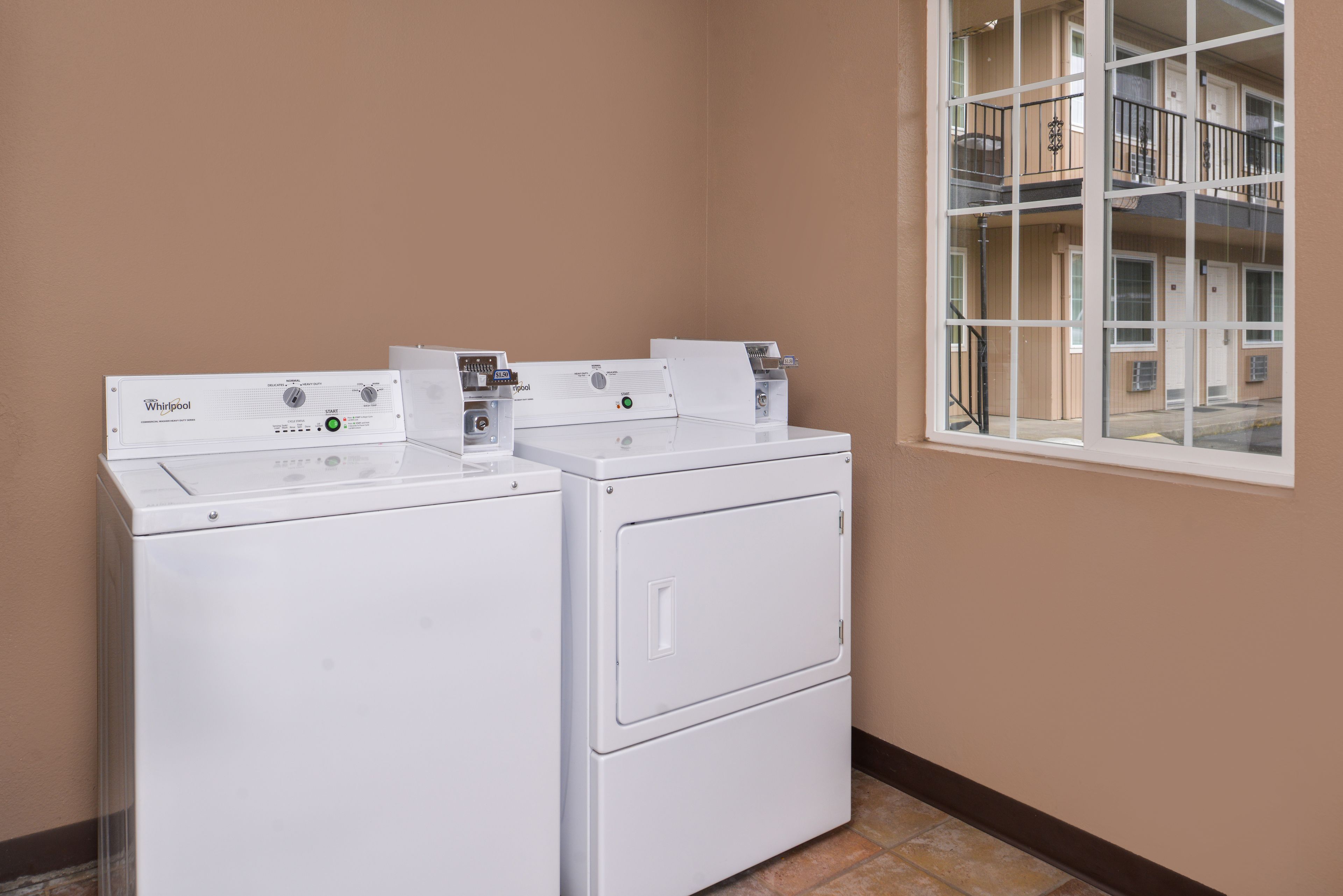 A laundry room with two Whirlpool washers and dryers, a window with a view of a building, and a brown wall.