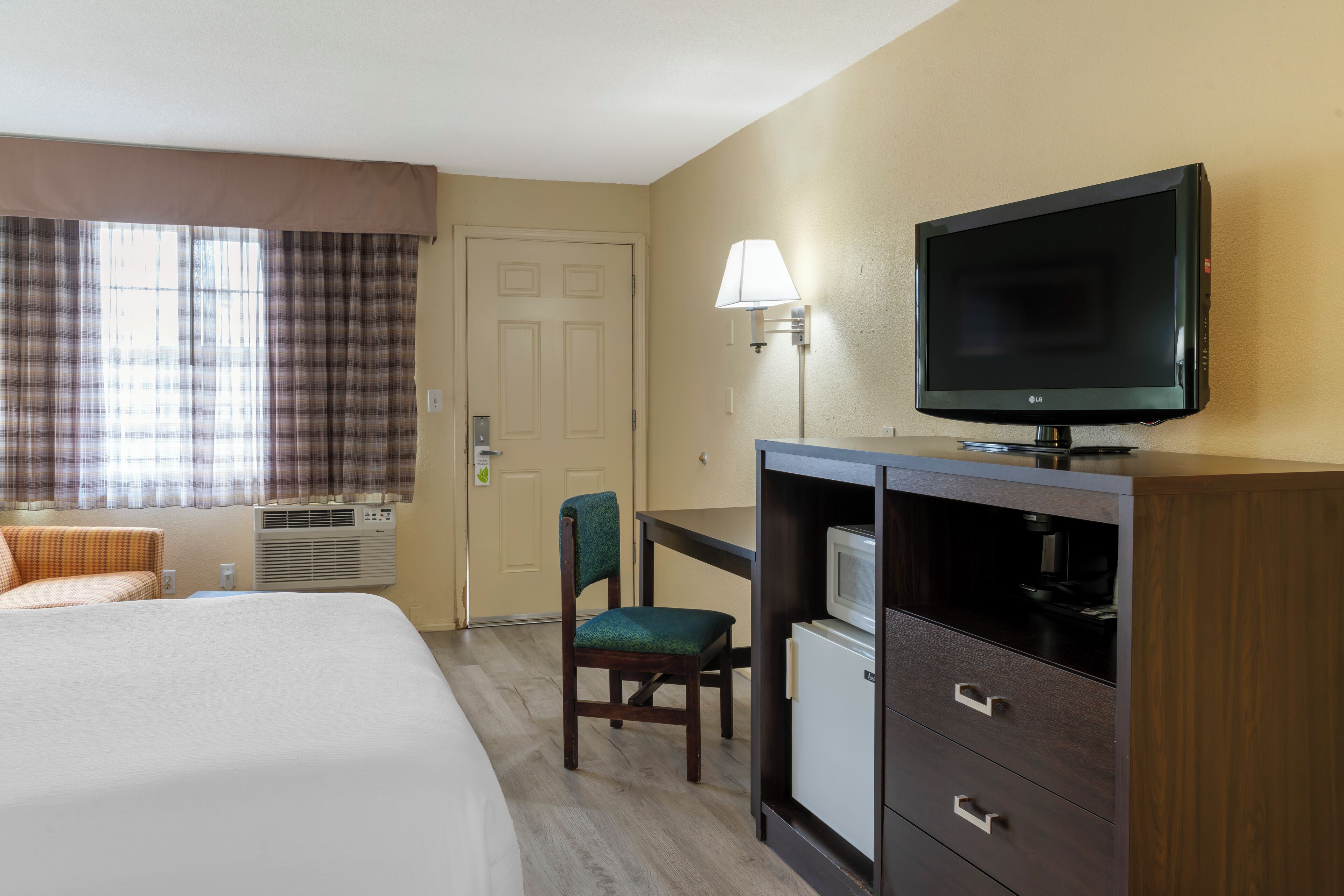 Habitación de hotel con cama, escritorio con televisor, silla, lámpara y puerta.