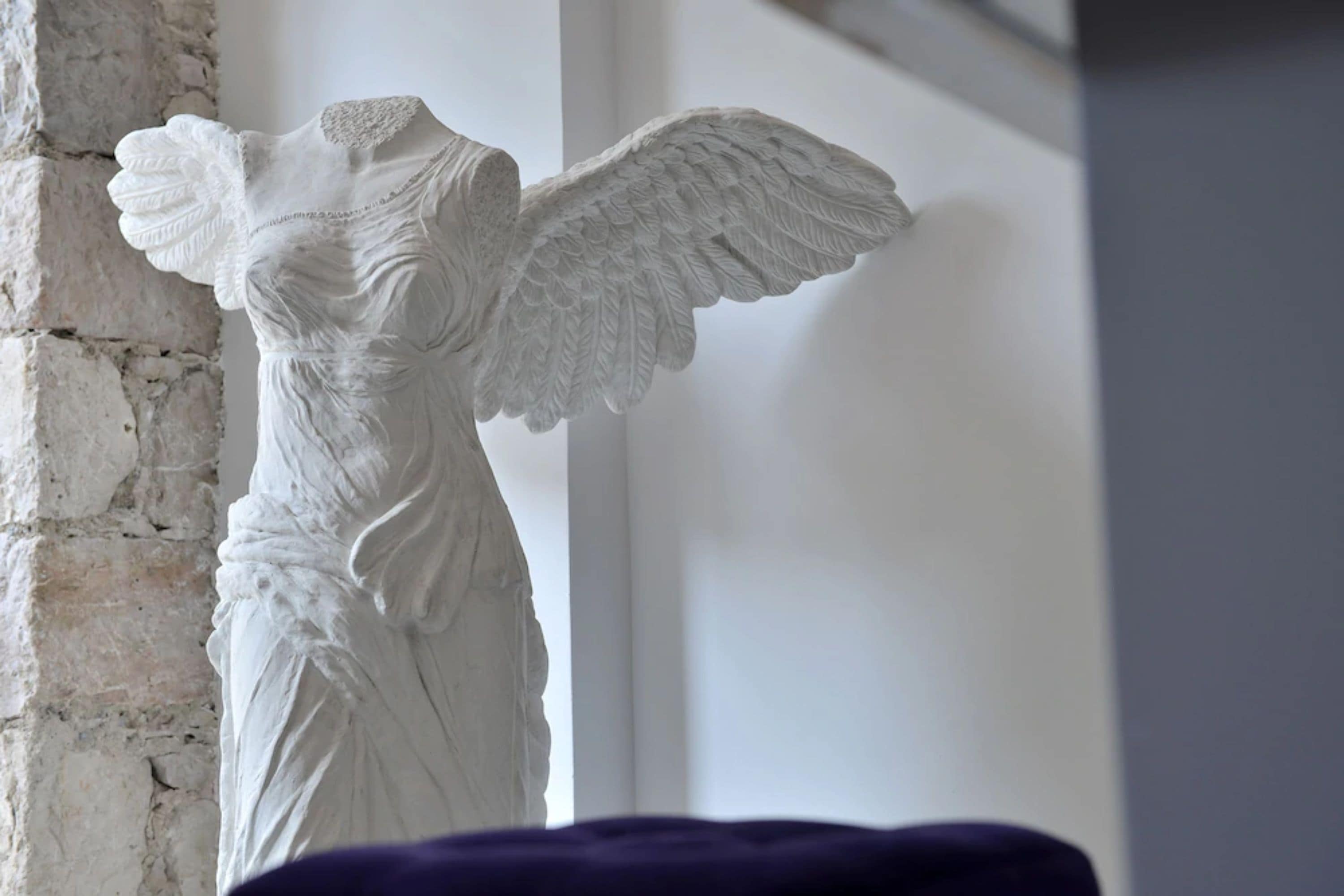 A white marble statue of an angel with outstretched wings, positioned against a white wall with a brick texture on the left.