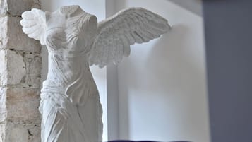 Estátua de mármore branco de um anjo com asas abertas, posicionada diante de uma parede branca com textura de tijolos à esquerda.