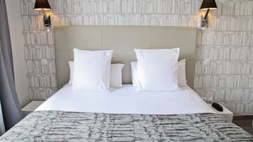 Una cama bien tendida con sábanas blancas y una manta decorativa. Hay dos lámparas fijadas a la pared a cada lado de la cama.