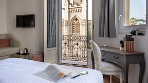 A hotel room with a bed, a desk, a chair, a television, and a view of a building through an open door. - Hôtel La Villa Nice Victor Hugo (Nice)