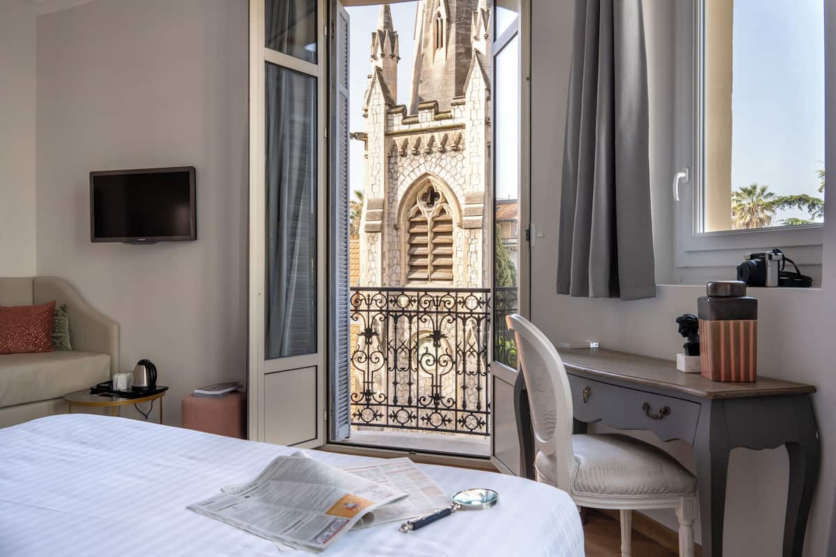 A hotel room with a bed, a desk, a chair, a television, and a view of a building through an open door.