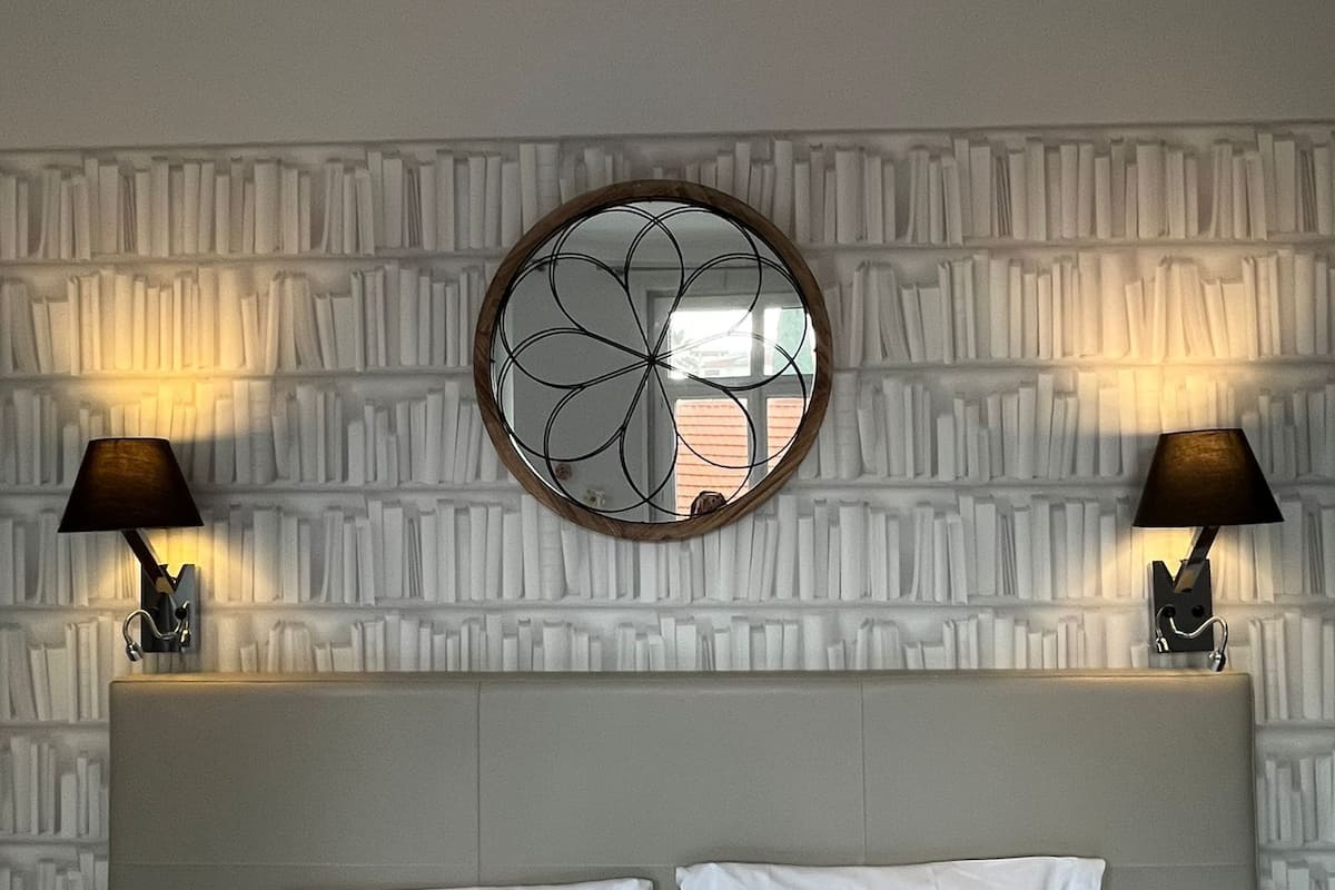 A hotel room with a large bed, two bedside lamps, a decorative wall panel, and a round mirror above the bed.