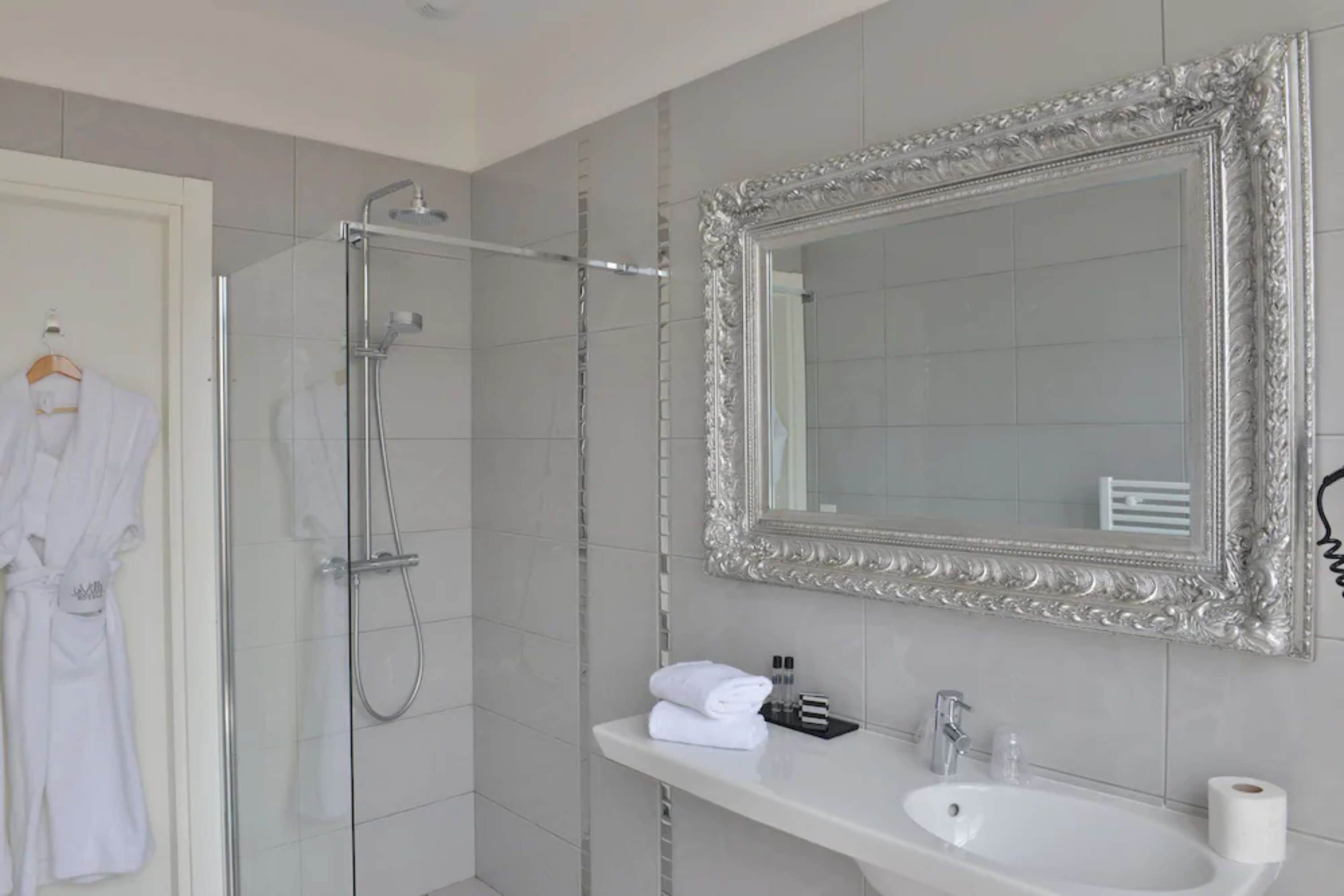 Un baño con cabina de ducha de vidrio, un lavabo blanco, un espejo grande y decorativo, y una bata blanca colgada en la pared.