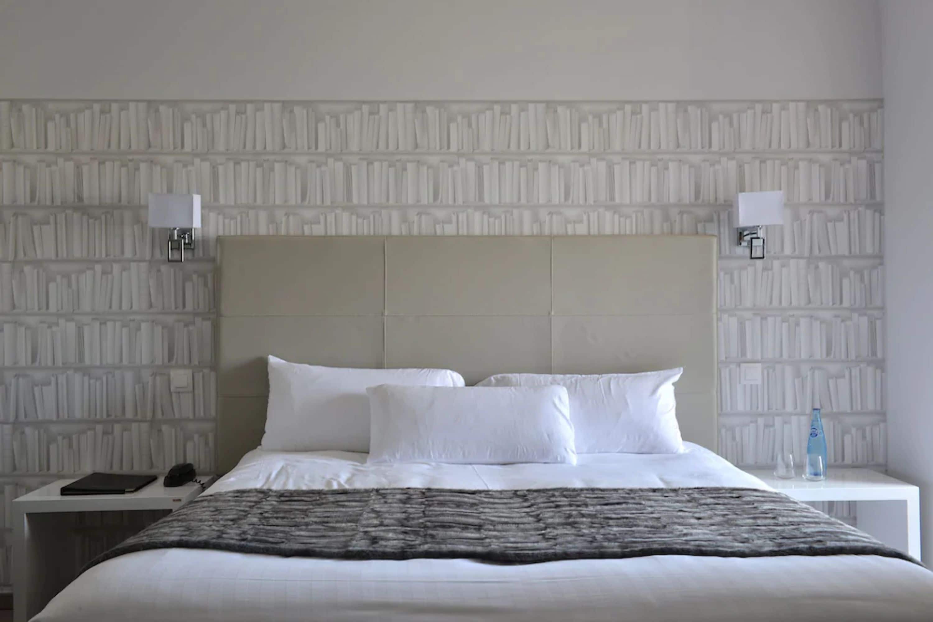 A neatly made bed with white linens and a gray throw blanket, a bedside table with a lamp, and a wall with a textured pattern.
