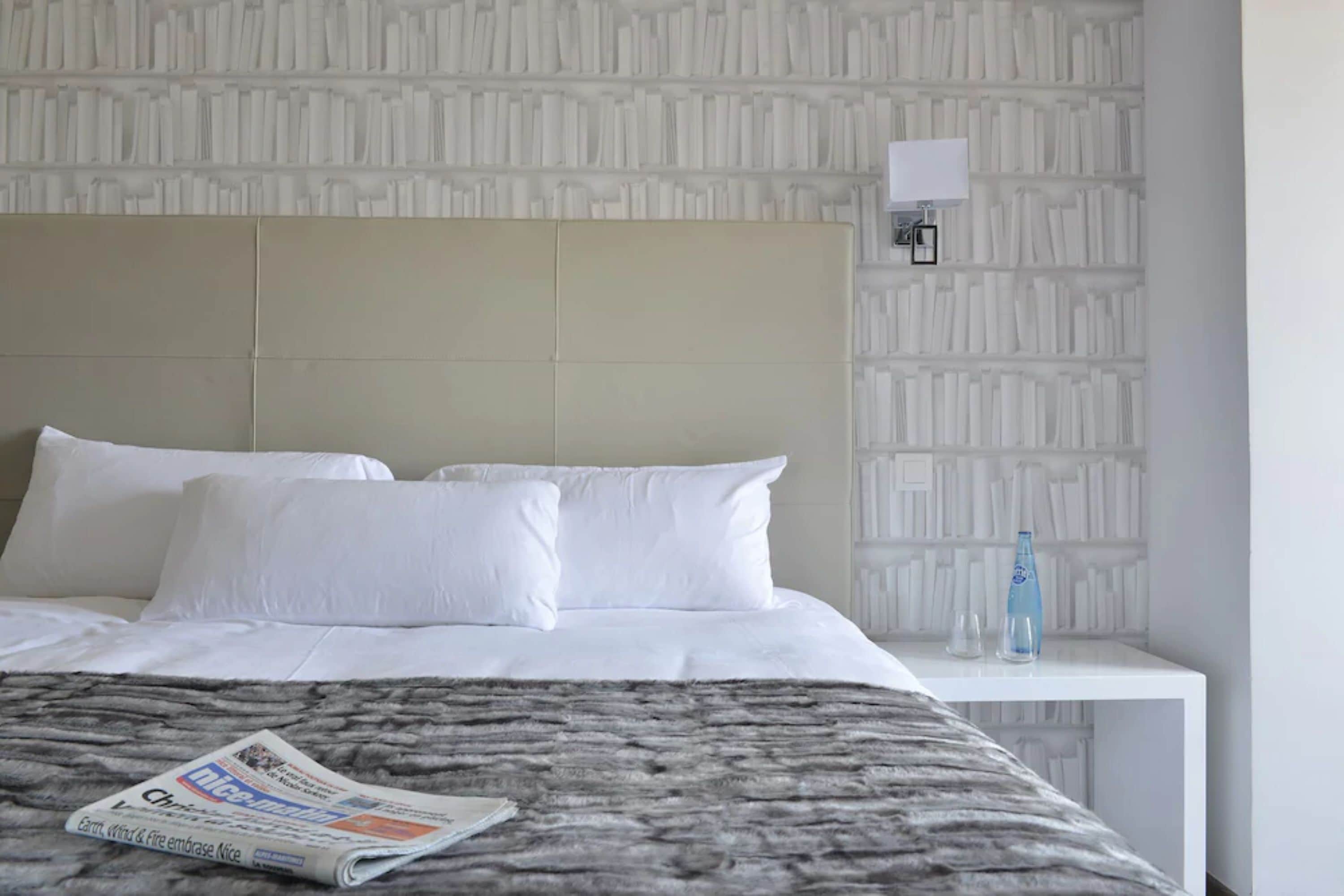 Una cama bien hecha con sábanas blancas, un edredón gris con textura y un periódico doblado sobre la cama.
