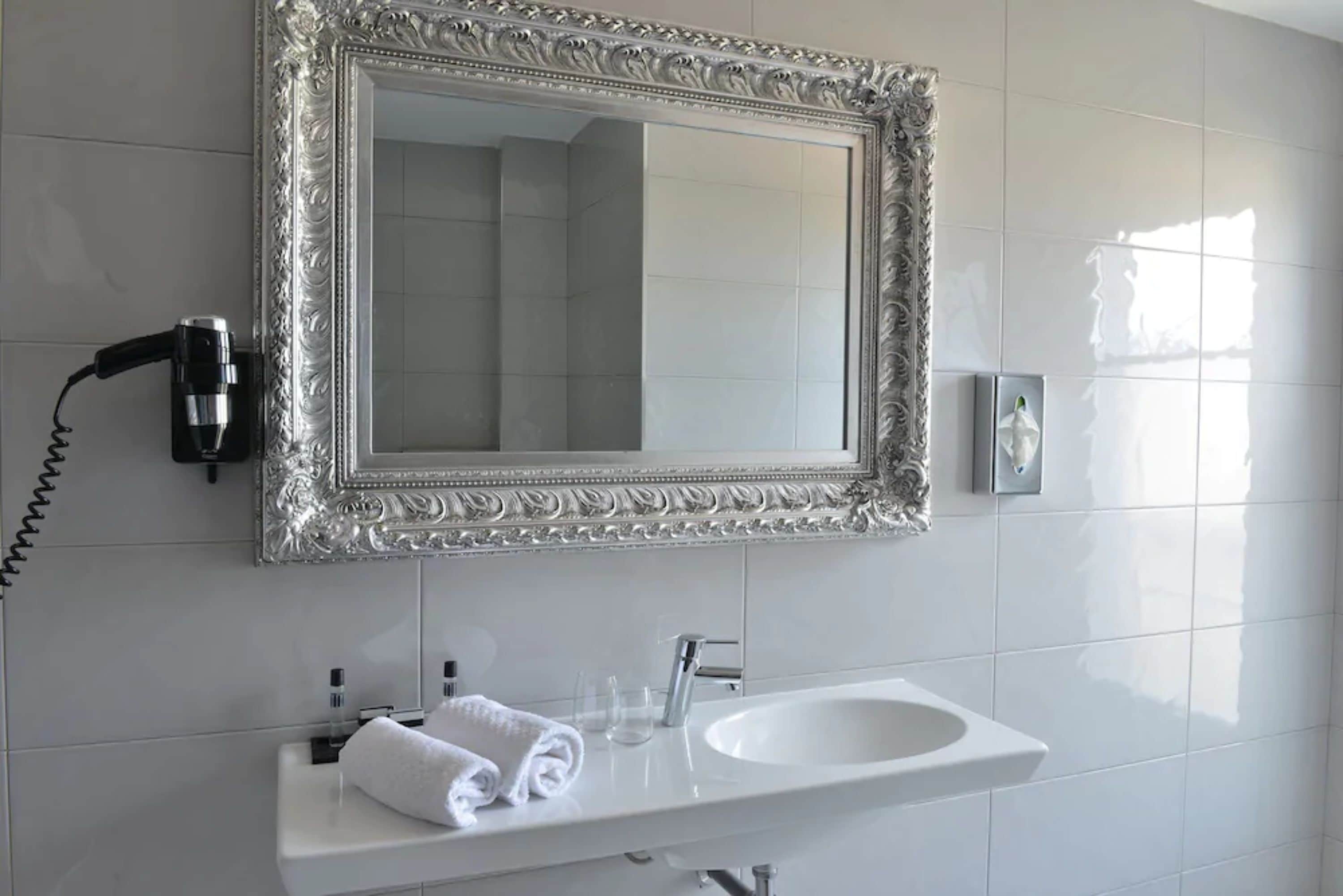 Un baño con lavamanos blanco, espejo con marco decorativo, secador de pelo y dispensador de pañuelos.