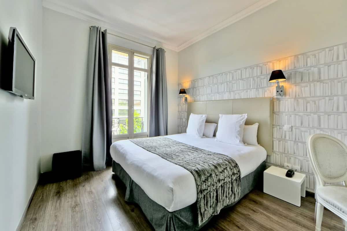 A hotel room with a large bed, a flat-screen TV, a bedside table with a lamp, and a view of greenery through the window.