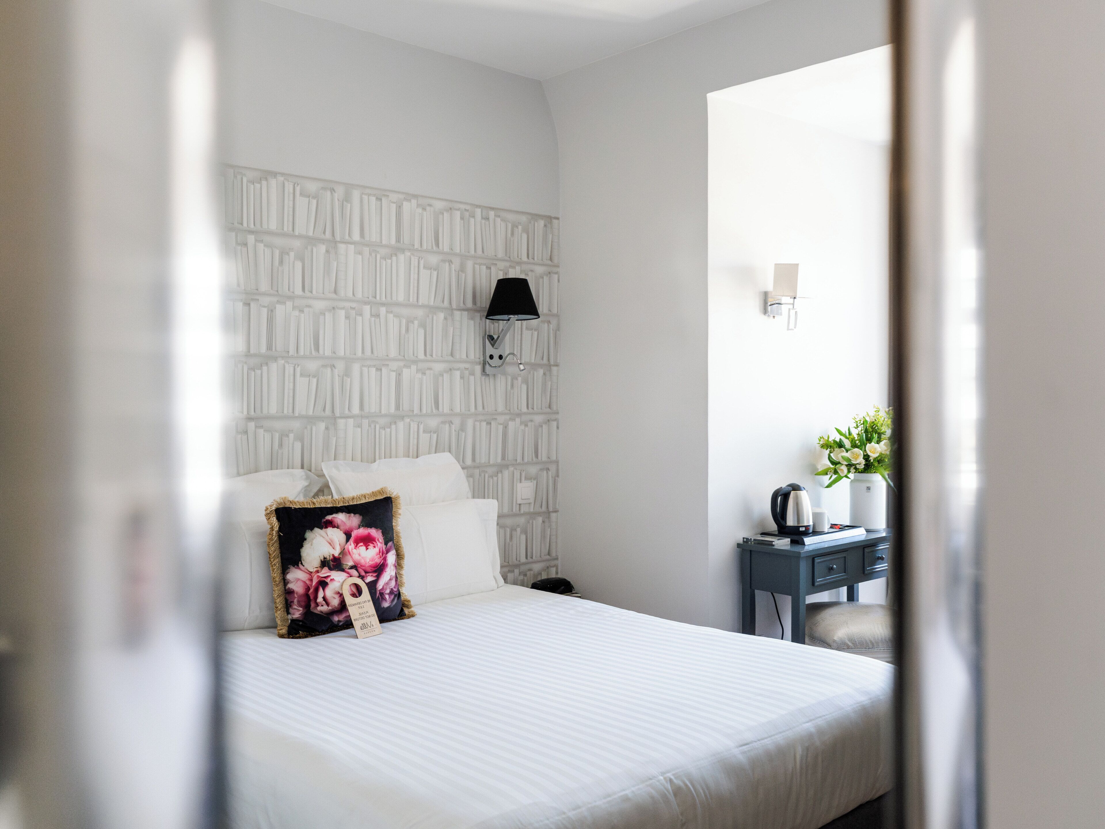 Une chambre d’hôtel avec un lit, une table de chevet, une lampe fixée au mur et une petite table sur laquelle est posé un vase de fleurs.