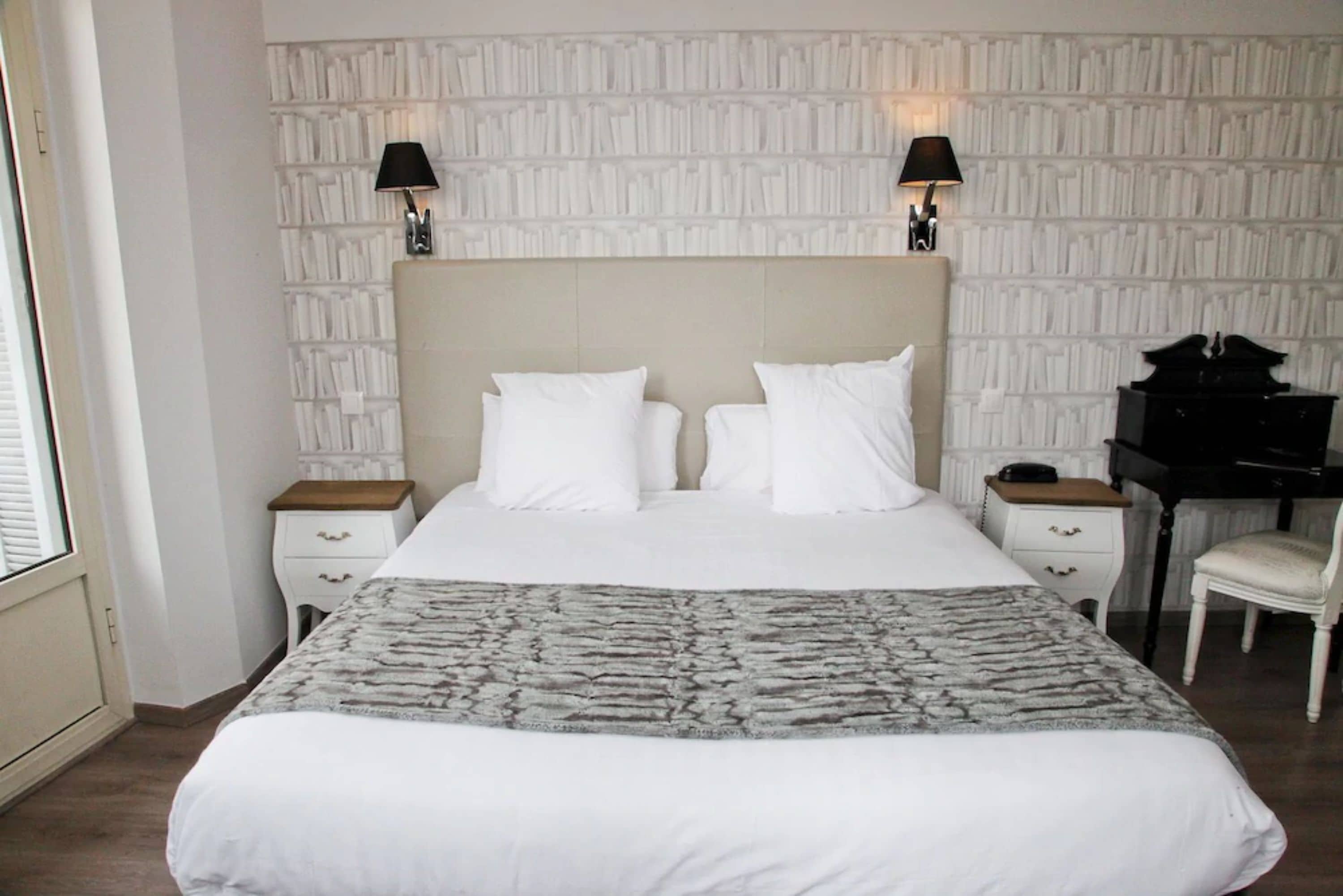 A neatly made bed with white linens, two bedside tables with lamps, a desk with a chair, and a patterned wall.