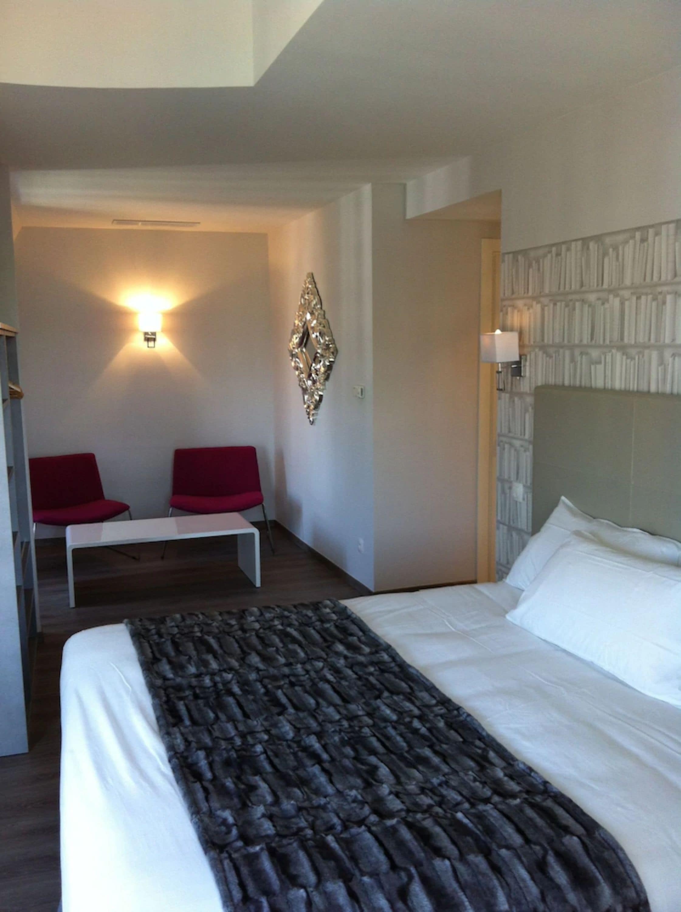 A hotel room with a bed, a bench, two red chairs, a wall-mounted light, and a decorative wall piece.