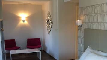 Habitación de hotel con cama, un banco, dos sillas rojas, una lámpara de pared y un adorno mural.