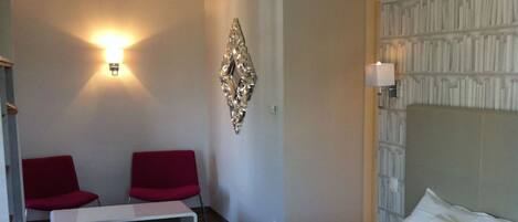 A hotel room with a bed, a bench, two red chairs, a wall-mounted light, and a decorative wall piece.