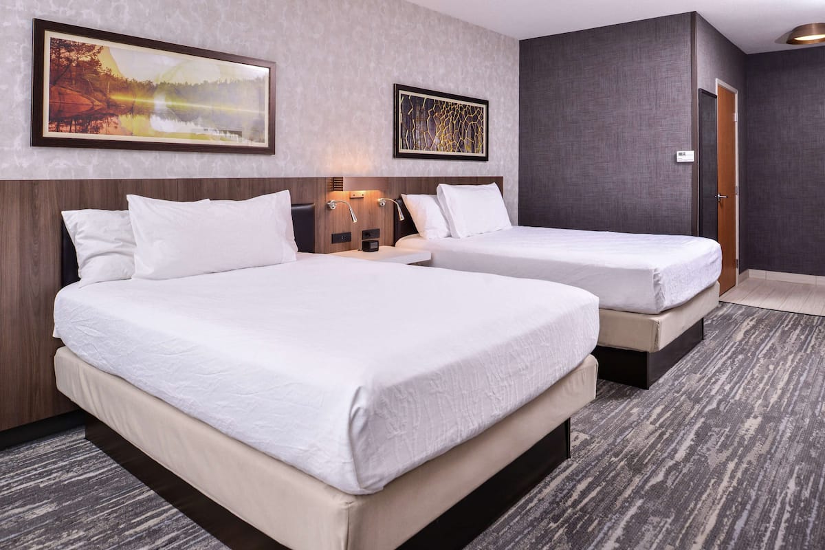 Quarto de hotel com duas camas, cabeceira, mesa de cabeceira e luminária fixada na parede.