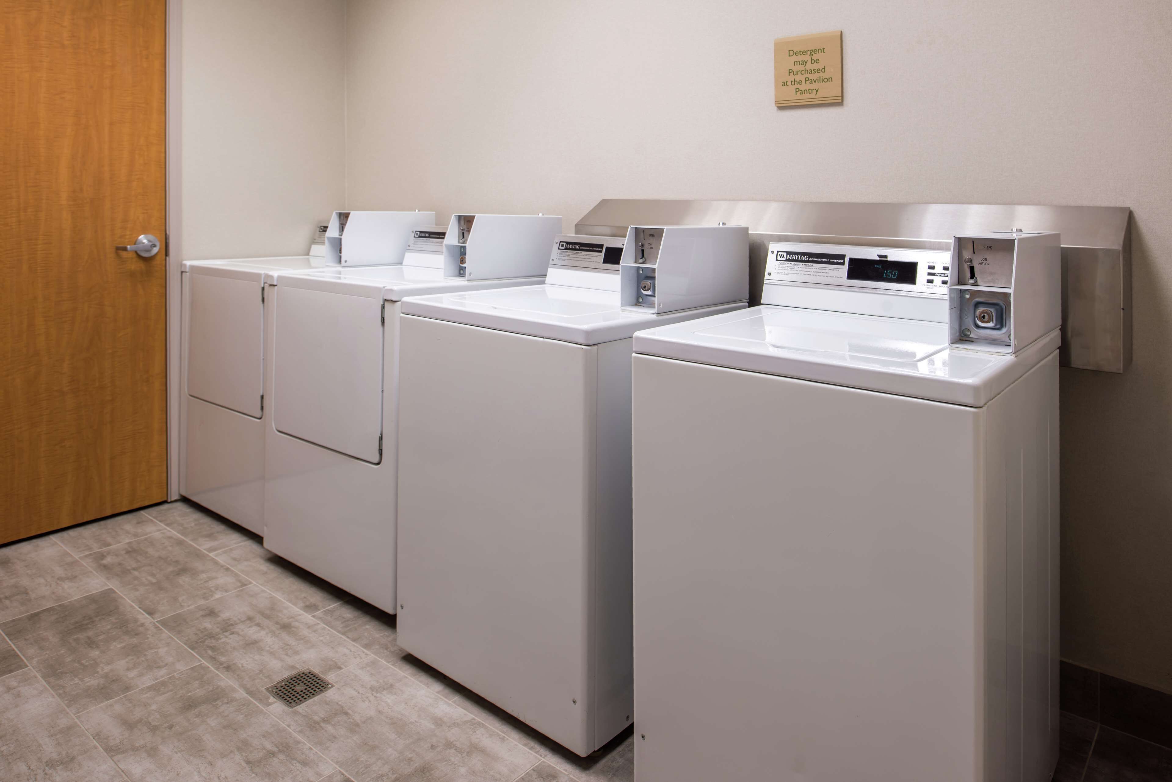 A laundry area with four white front-loading washers and dryers, a wooden door, and a sign indicating the location.