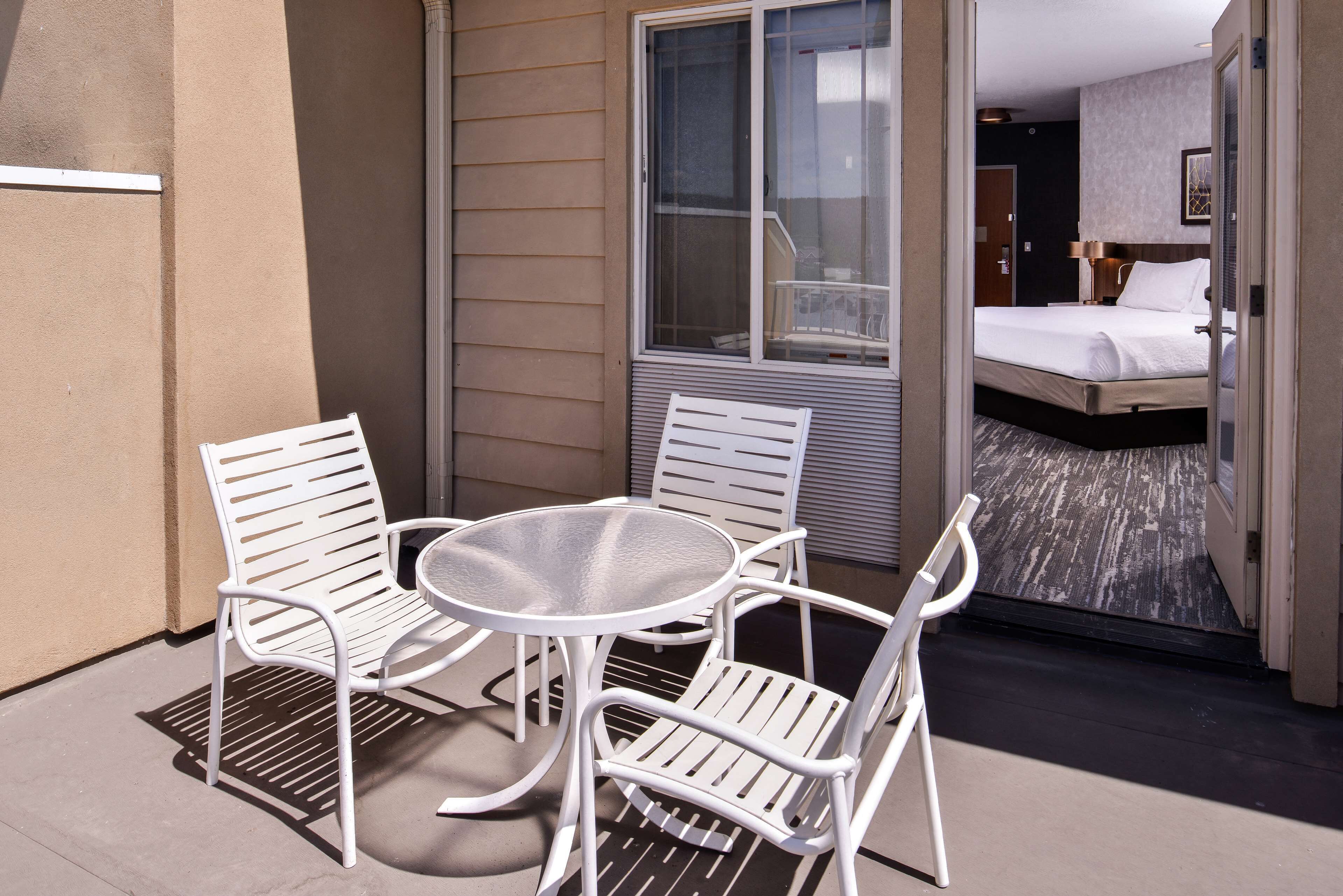 Un balcon avec une table et quatre chaises, une porte-fenêtre coulissante donnant sur une chambre à coucher équipée d’un lit et d’une table de chevet.
