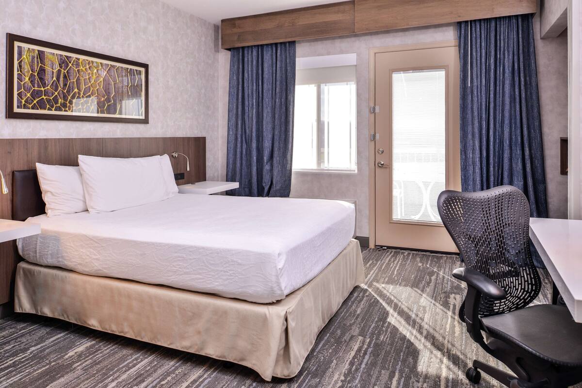 Quarto de hotel com cama, escrivaninha, cadeira, janela grande e porta.