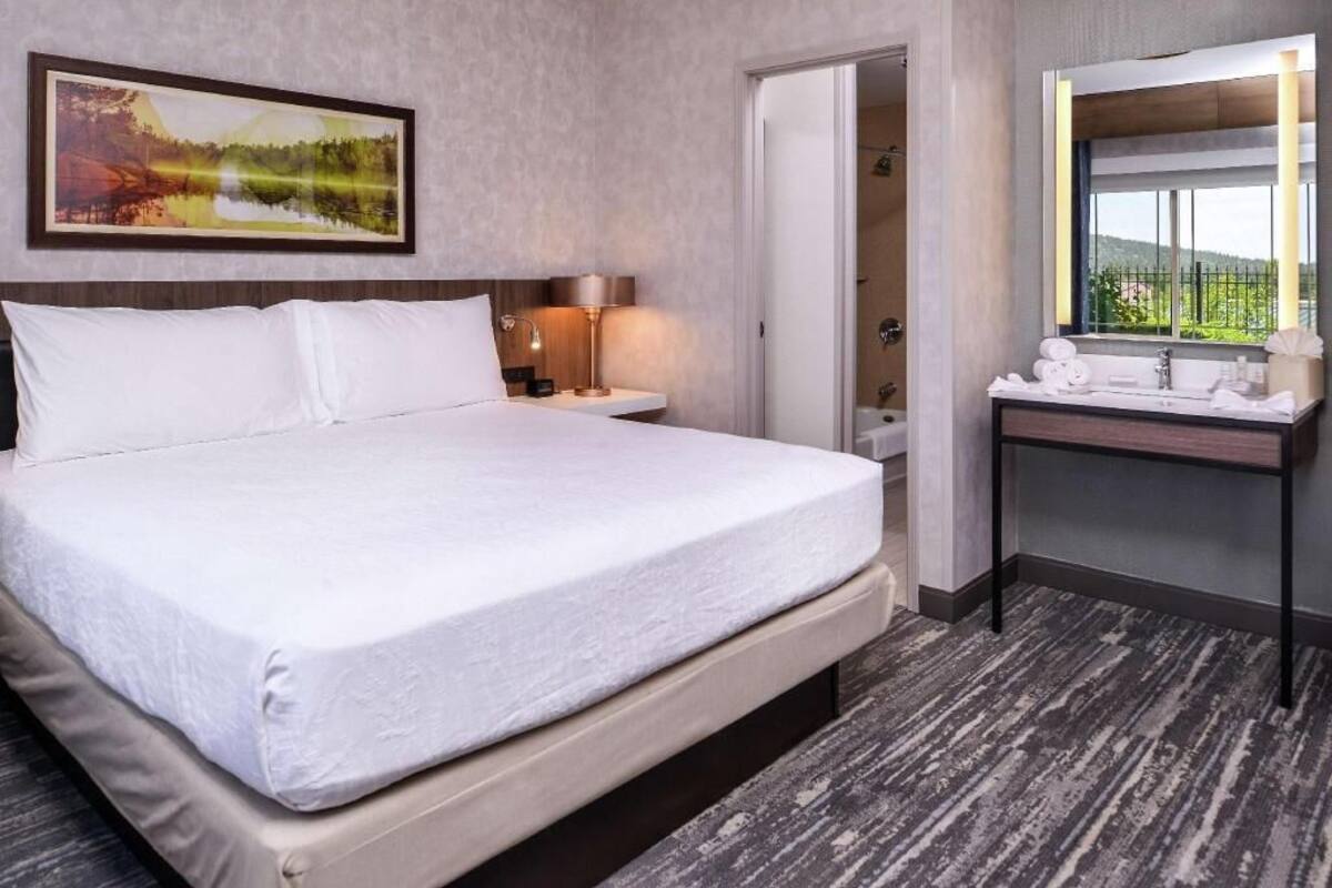Quarto de hotel com cama, mesa de cabeceira com abajur, banheiro com espelho e pia, e um quadro na parede.
