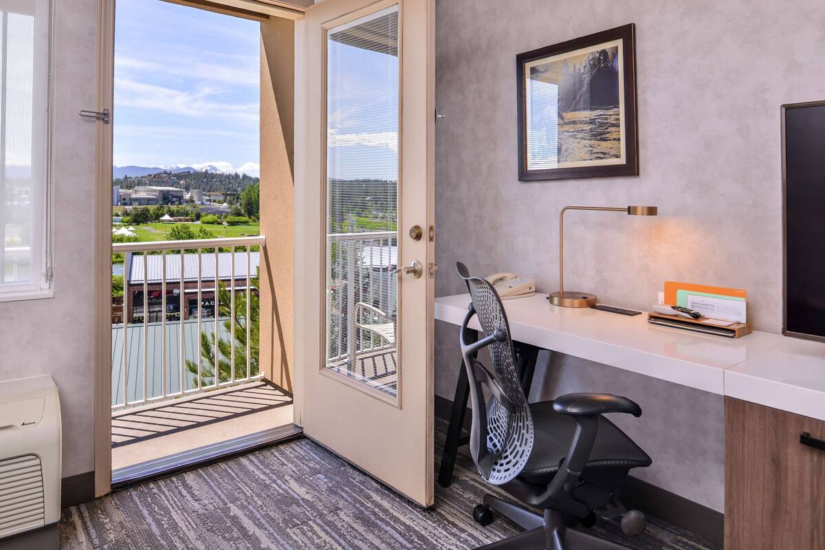 Um espaço de escritório moderno com uma mesa, cadeira e vista para uma varanda com paisagem urbana e montanhas ao fundo.