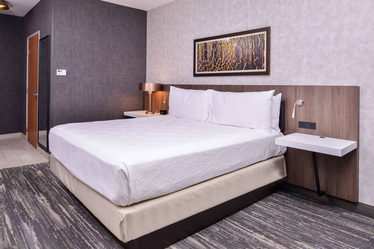 Quarto de hotel moderno com uma cama grande, mesas de cabeceira, uma obra de arte pendurada na parede e uma pequena mesa de cabeceira.