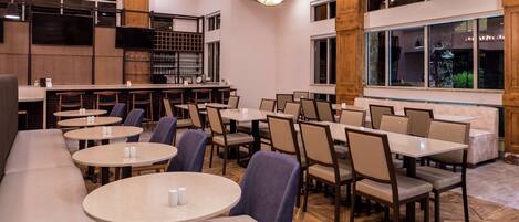 A restaurant with multiple round tables and chairs, a bar area, and large windows providing natural light.