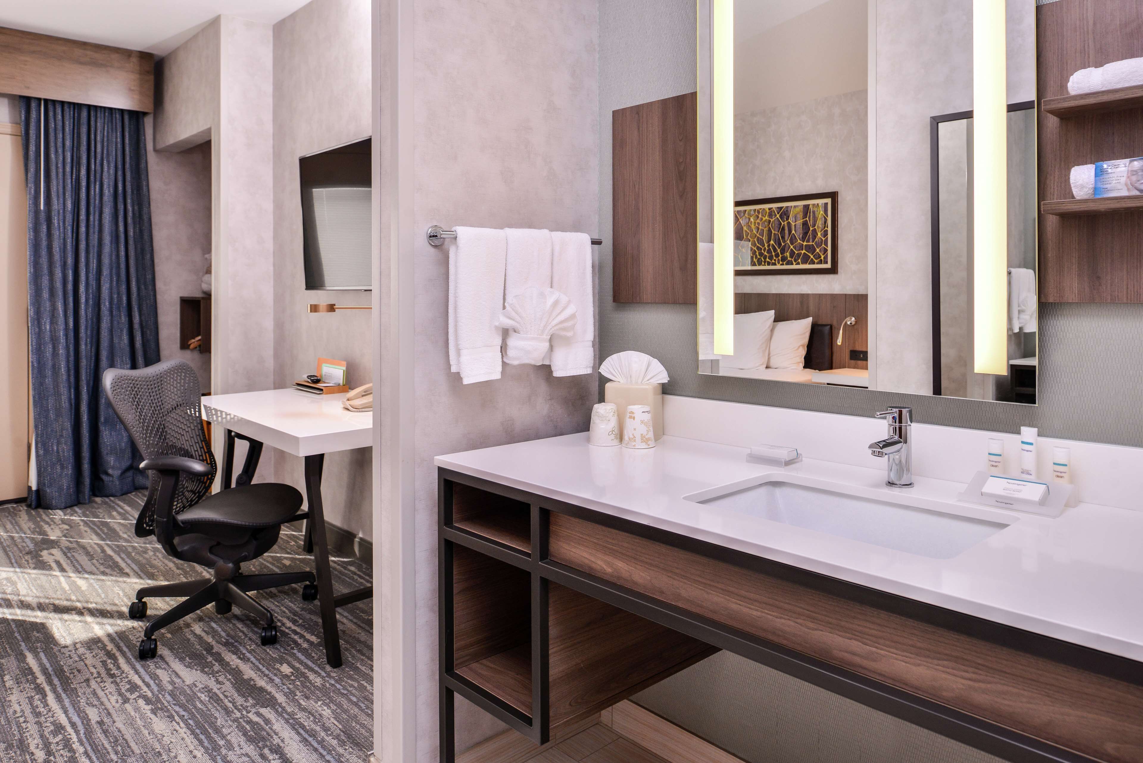 A modern hotel bathroom with a large sink, a mirror, a chair, and a desk.