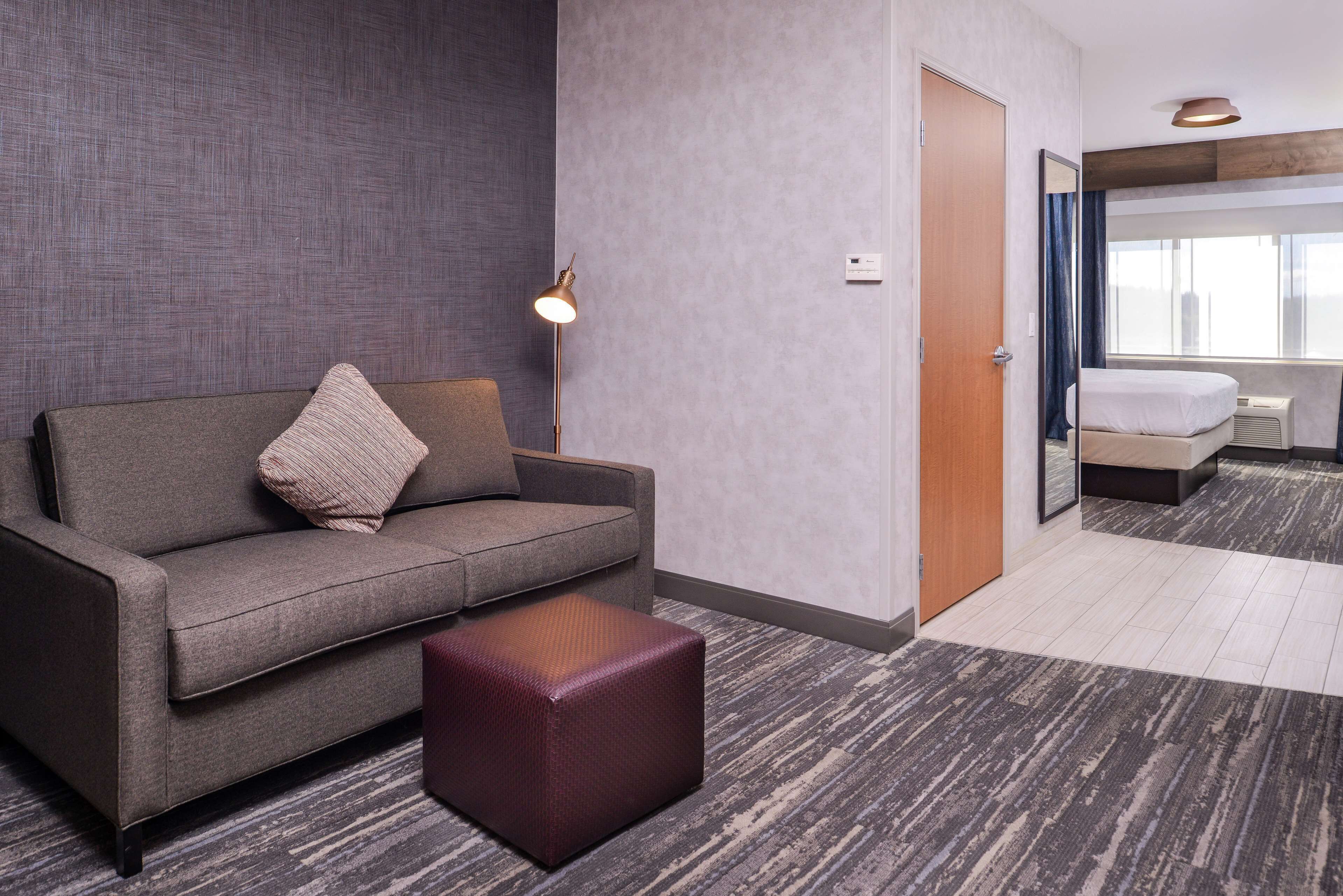 A hotel room with a grey sofa, a small ottoman, a floor lamp, and a bed with a white headboard.