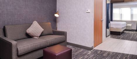 A hotel room with a grey sofa, a small ottoman, a floor lamp, and a bed with a white headboard.